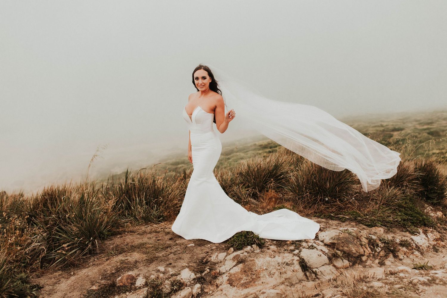 A bride in a form-fitting white dress poses dramatically with her veil flowing in the wind on a hilltop.