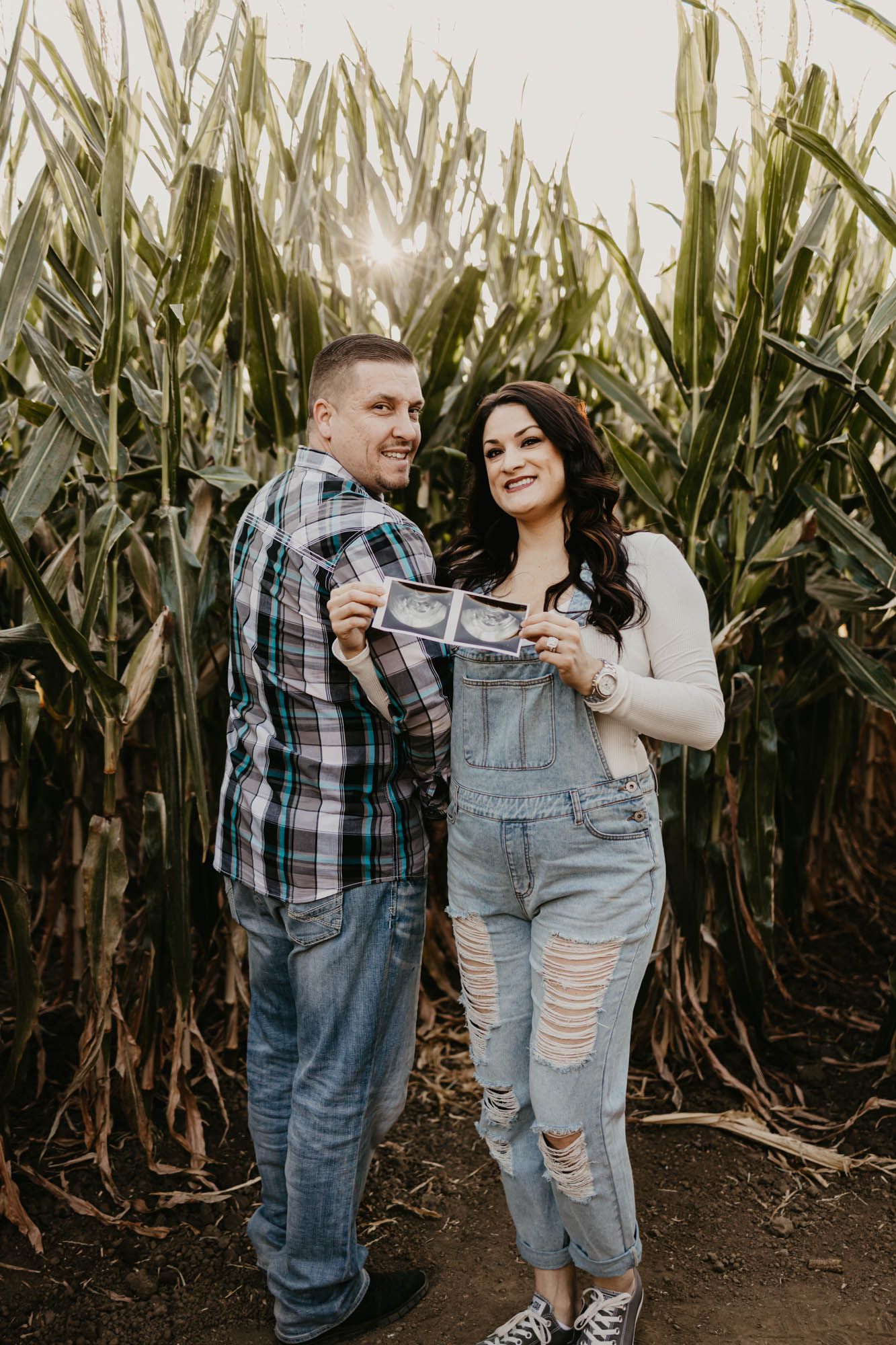 Expectant couple posing with sonogram photos against backdrop of tall corn plants.