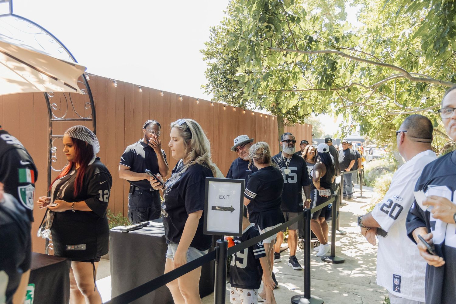Fans in Raiders jerseys queue outdoors at a sunny NFL game day event near a rustic wooden structure.