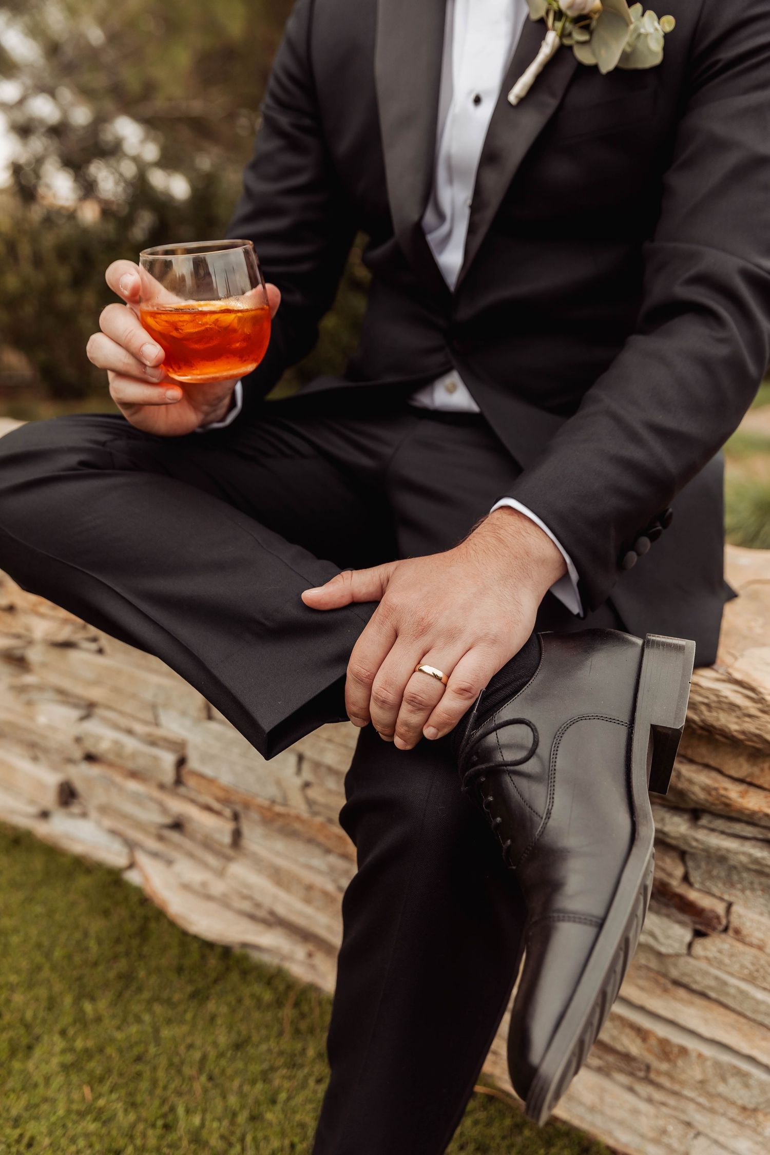 Groom in black tuxedo holding cocktail while sitting on stone wall.
