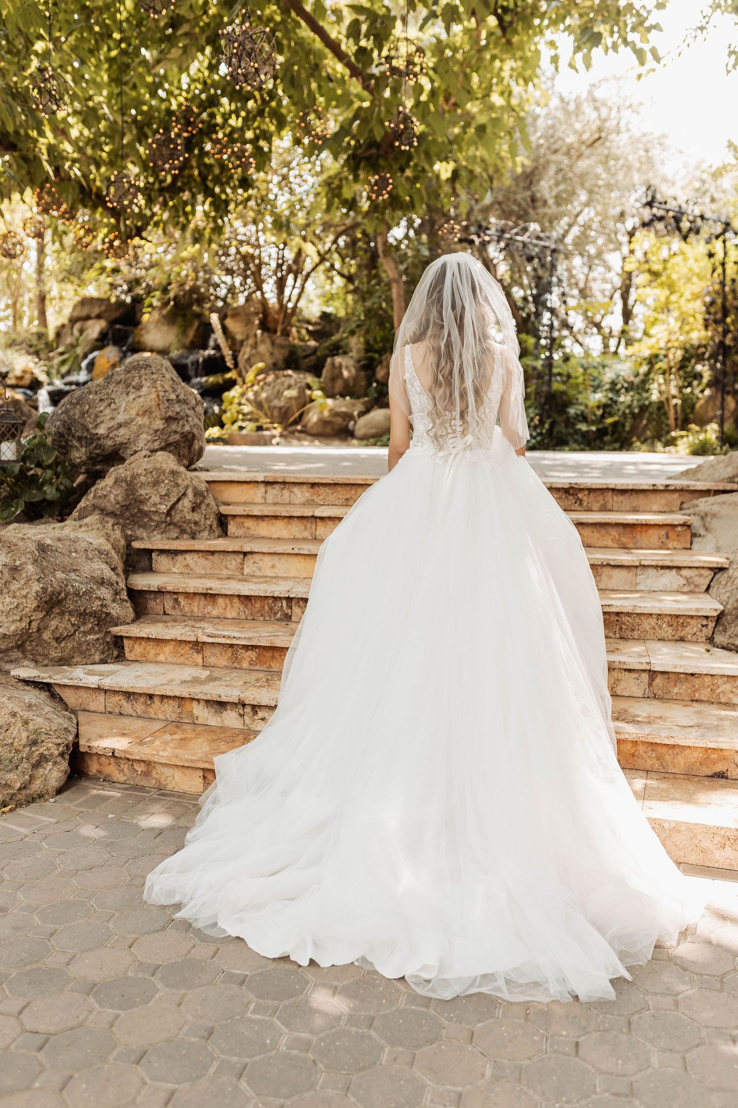 Back view of a wedding gown with cathedral train on stone garden steps surrounded by greenery.