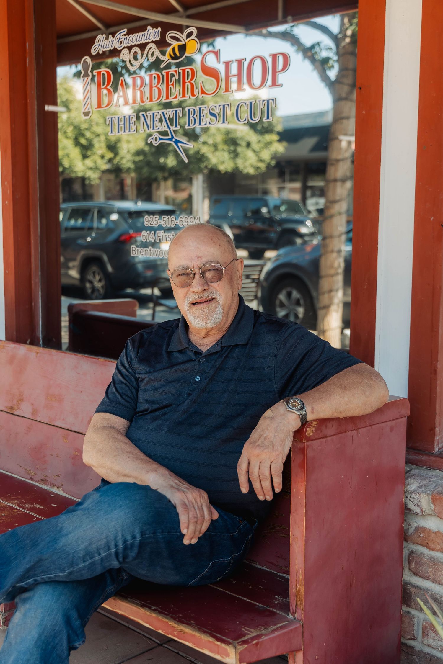 Portrait of someone in navy polo and jeans relaxing on a red bench outside a barber shop.