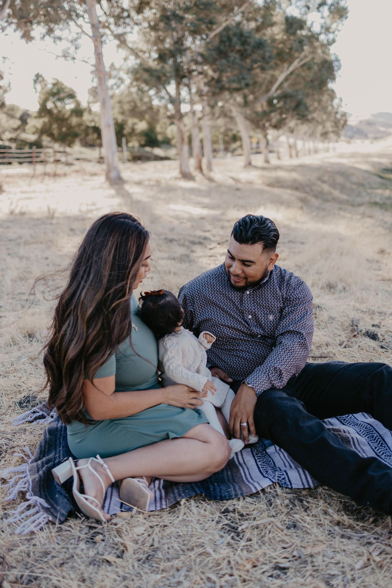 A young couple sits on a blanket outdoors, lovingly gazing at their newborn baby amid a serene park setting.
