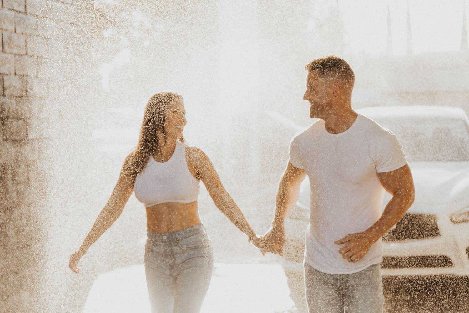 A couple in white tops and jeans hold hands while walking in soft sunlit setting.