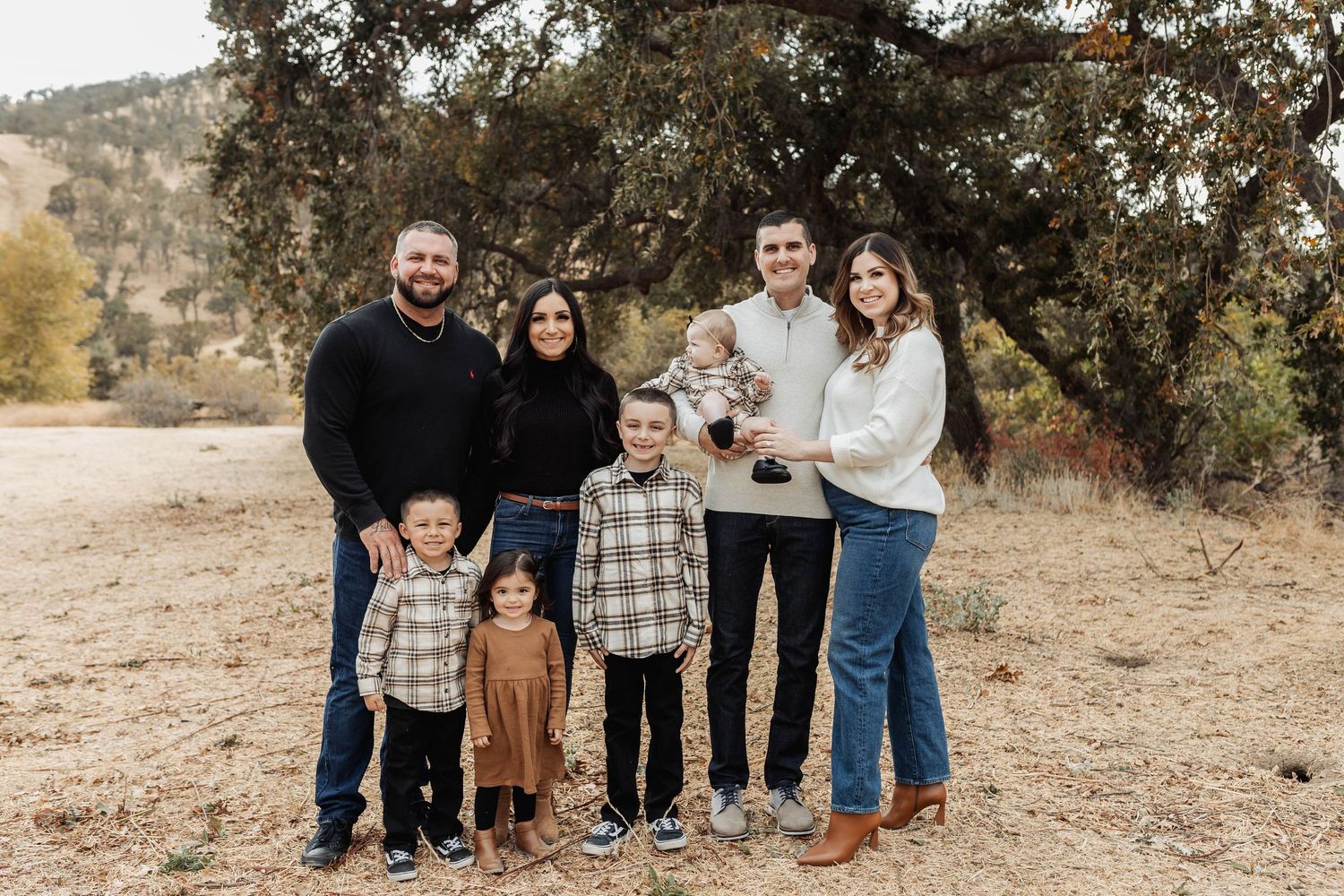 Family portrait of two adults and four children smiling outdoors in a natural setting with trees and dry grass.