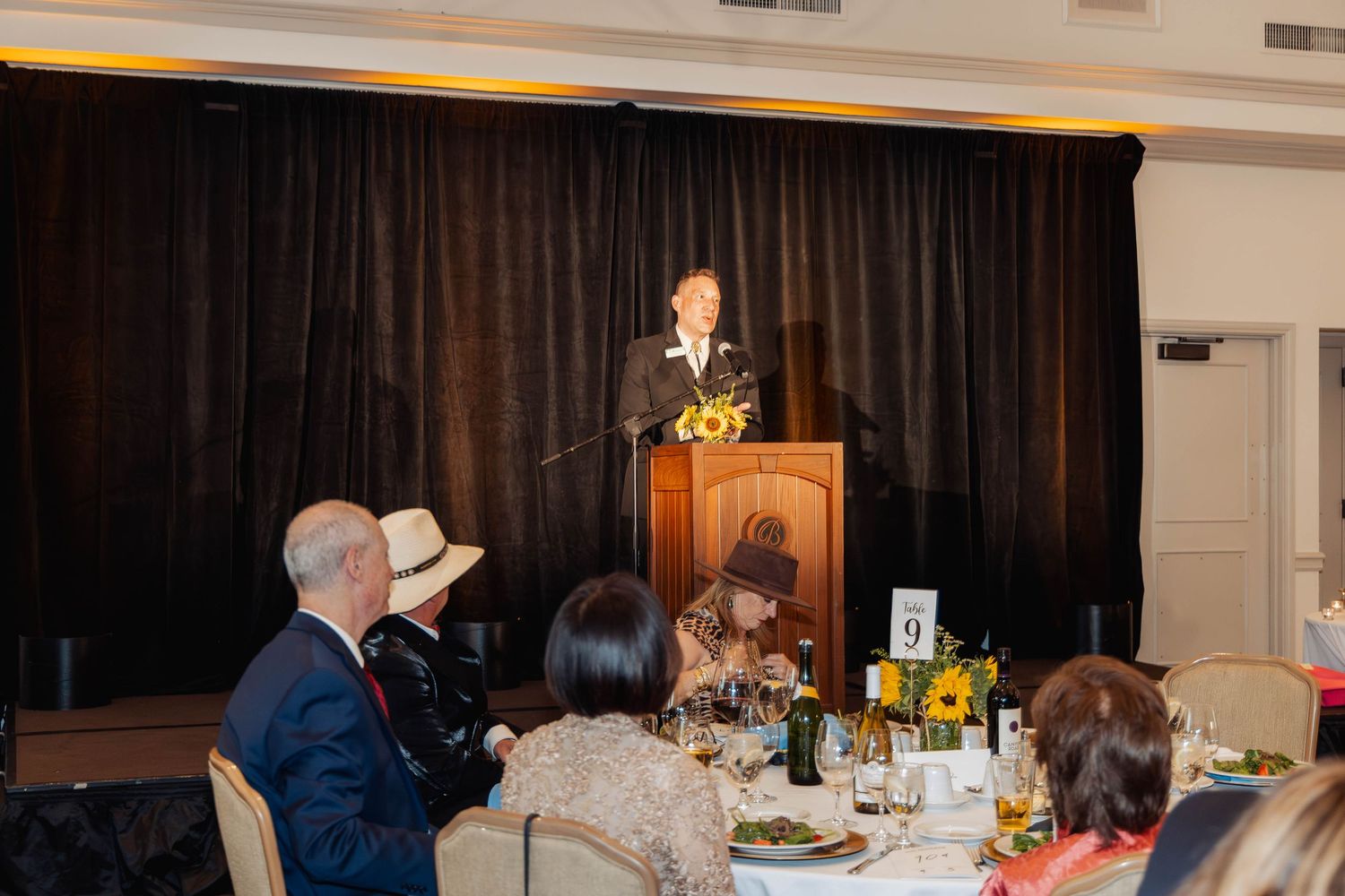 A speaker addresses guests at a formal gala dinner from a sunflower-decorated podium on a staged platform.