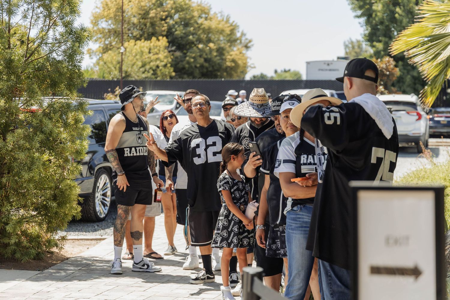 Raiders fans in black jerseys queue outdoors on a sunny day near an exit sign at a football event.