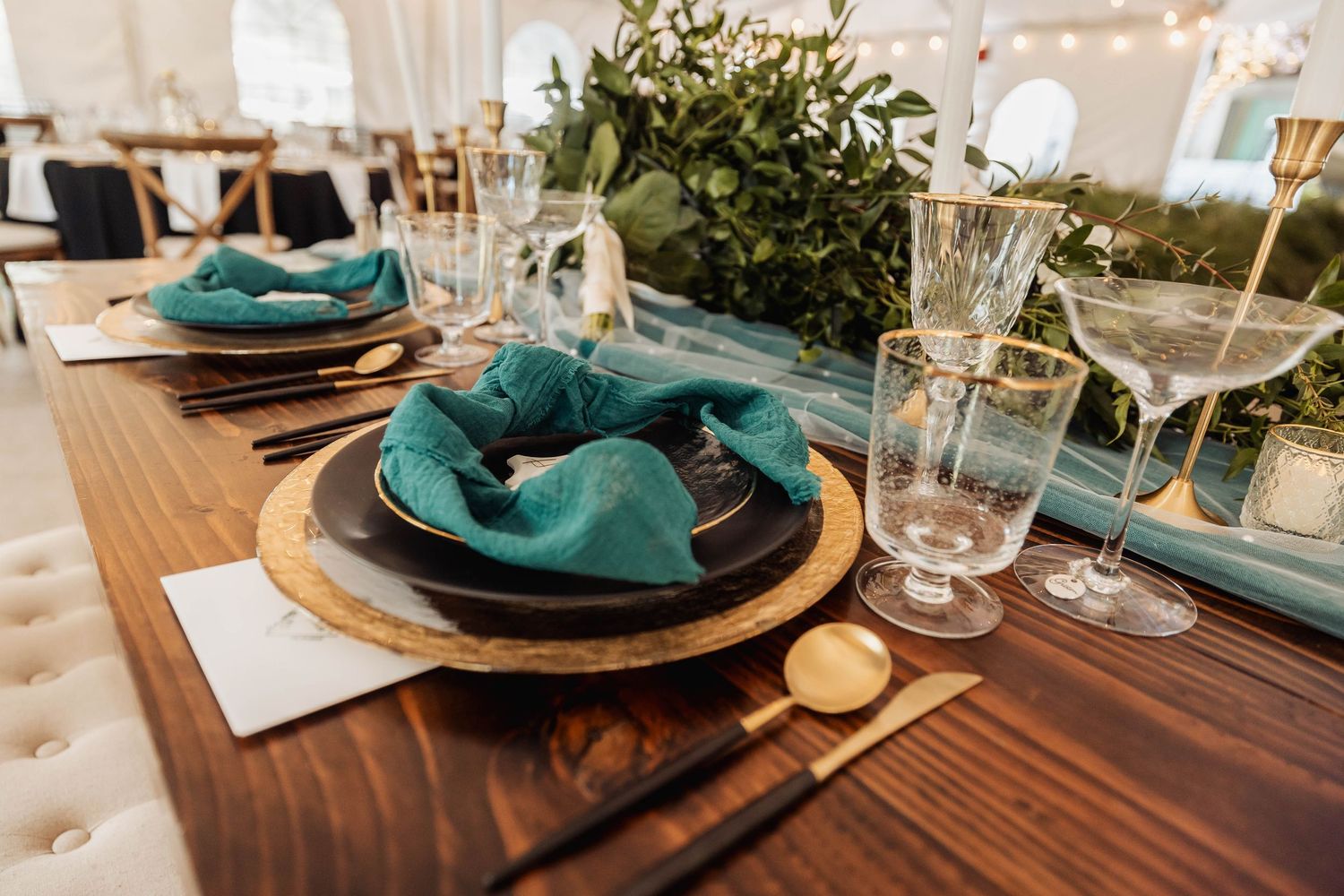 Wedding reception table setting with teal napkins, wooden tables, and string lights overhead creating an elegant ambiance.