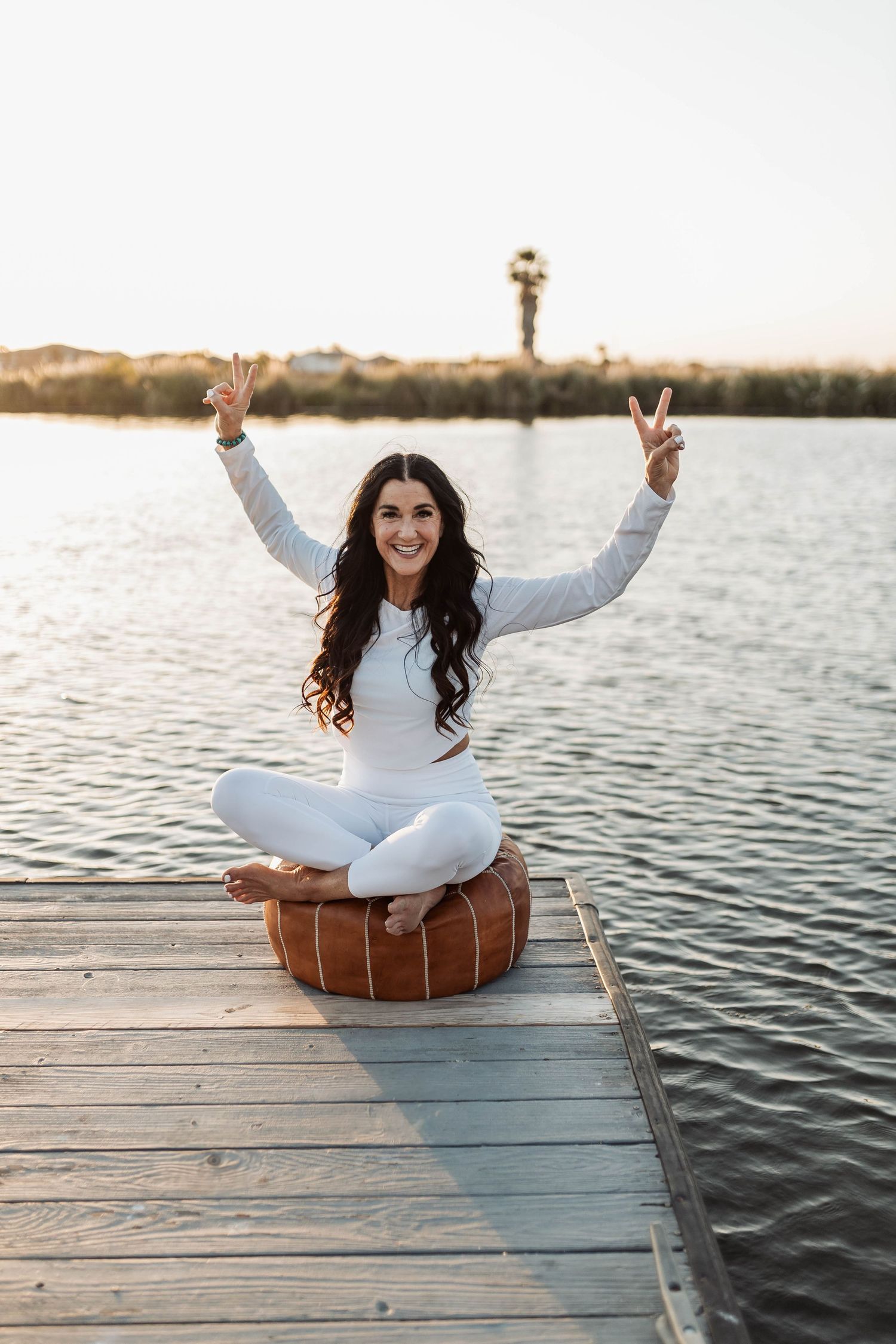Woman in white outfit making peace signs while sitting on dock by the water.