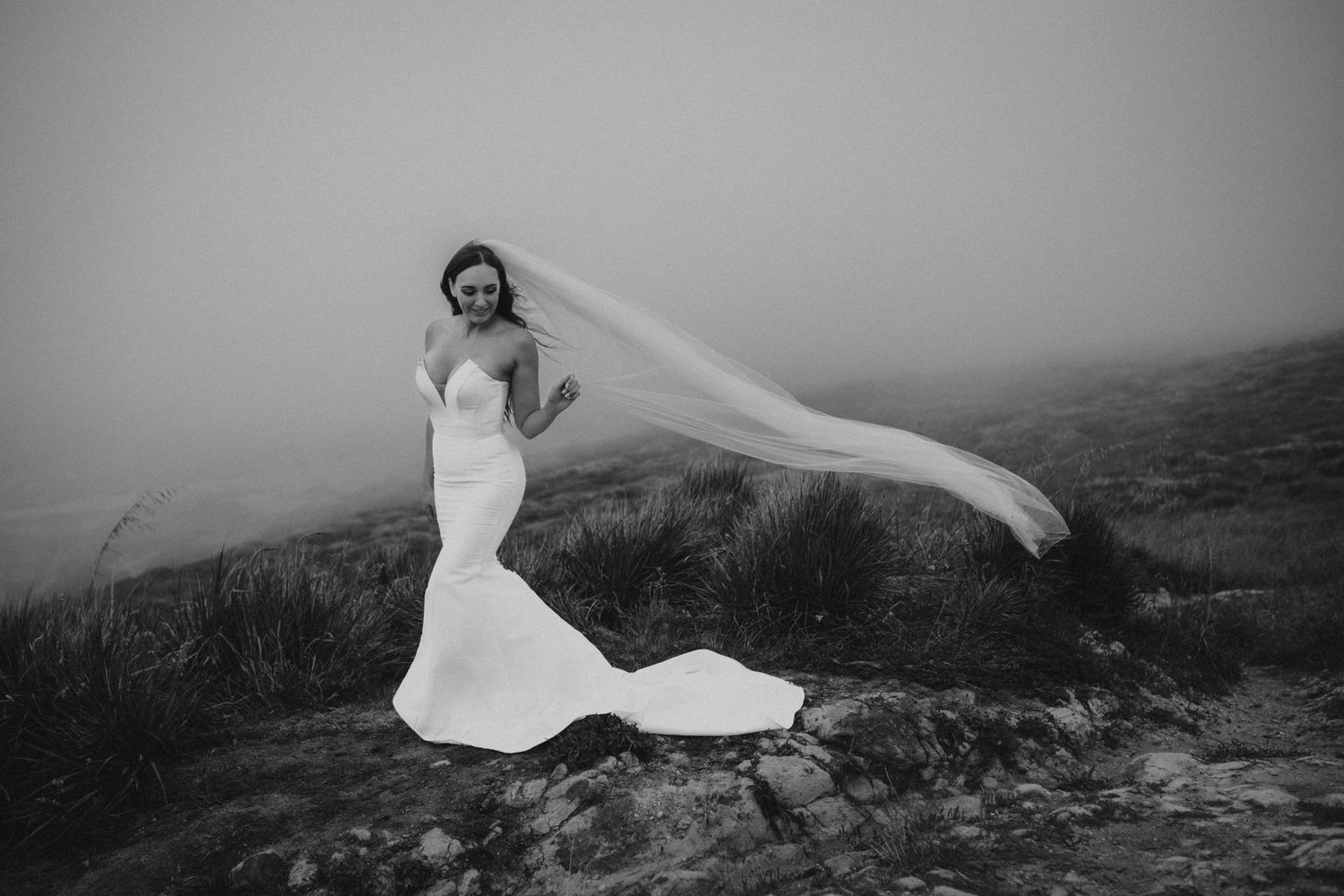 A black and white photo of a bride in a fitted wedding dress with flowing veil on a windswept hillside.
