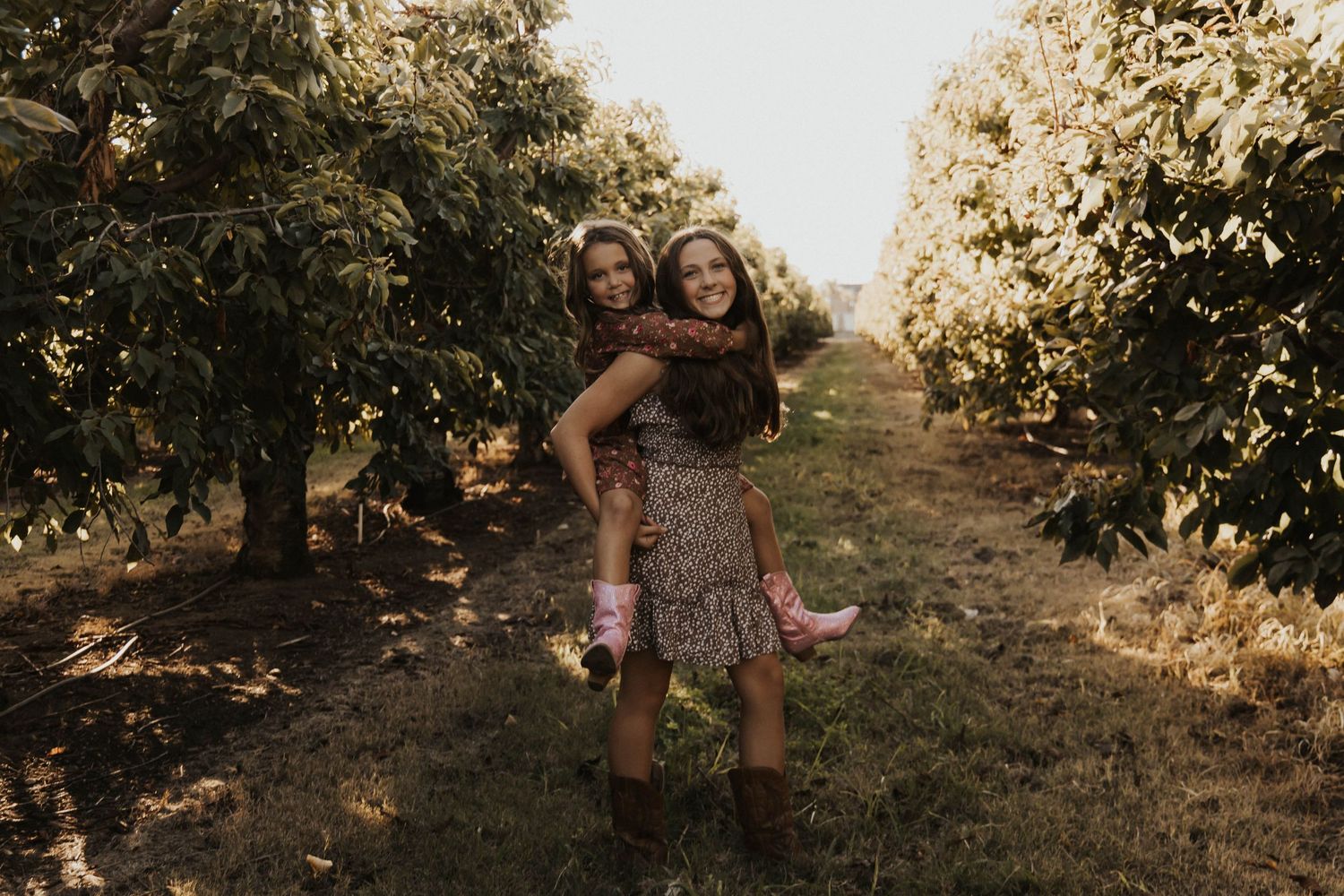 Two girls in floral dresses share a piggyback ride through a sunlit orchard with lush green trees.