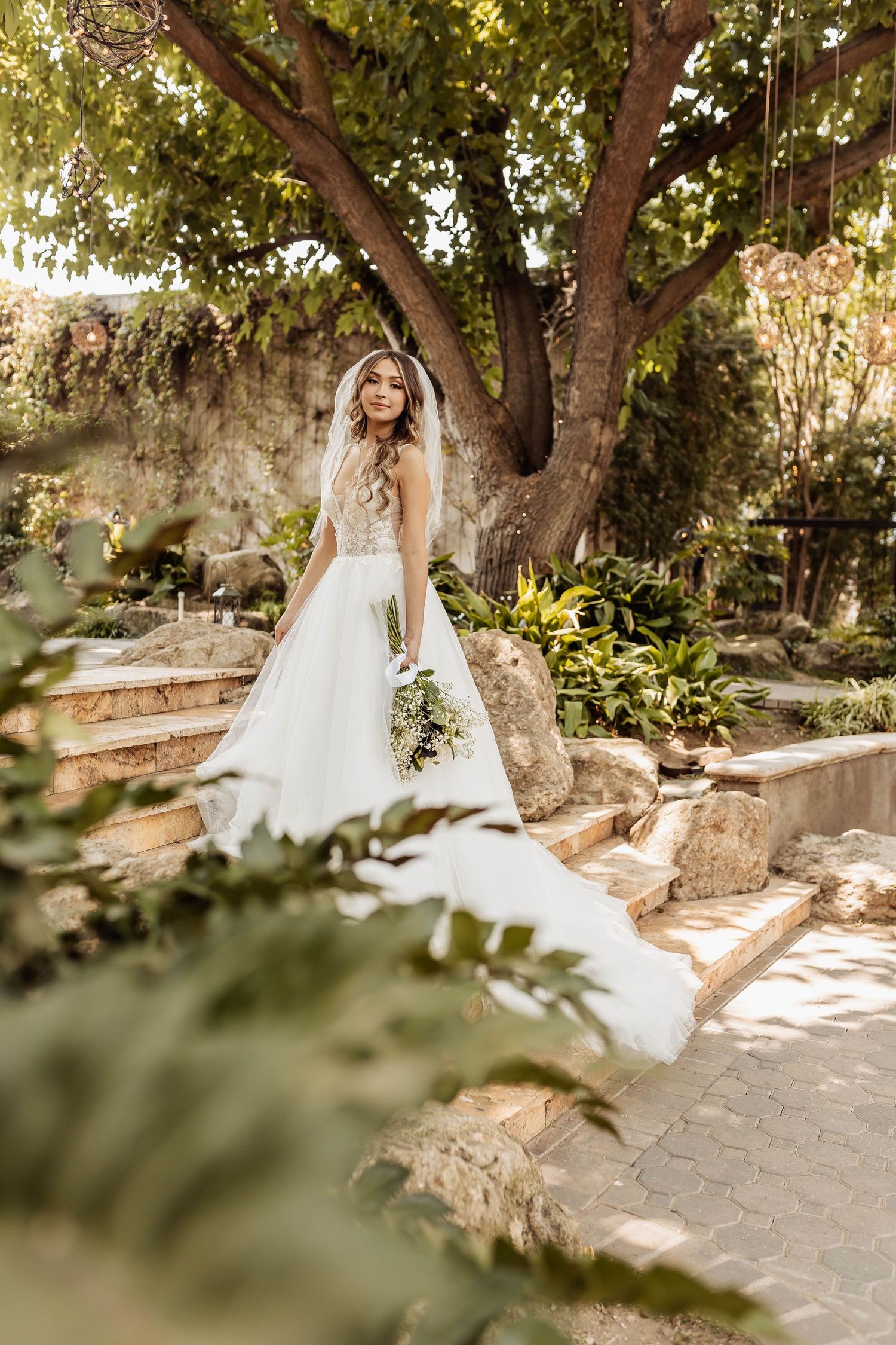 Ethereal bridal portrait series in a sunlit garden with flowing white wedding dress against stone walls and trees.