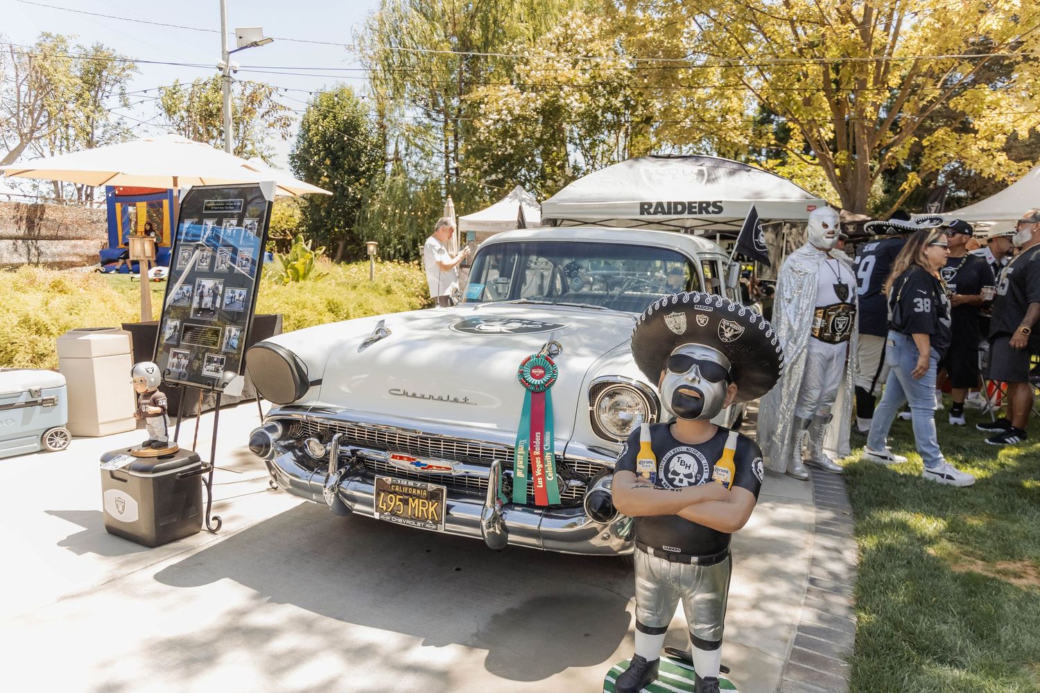Raiders fan in costume poses beside a decorated vintage white car at an outdoor tailgate party.