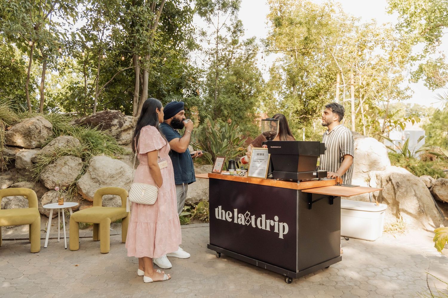 Customers ordering coffee at The Last Drip outdoor mobile coffee bar surrounded by lush greenery.