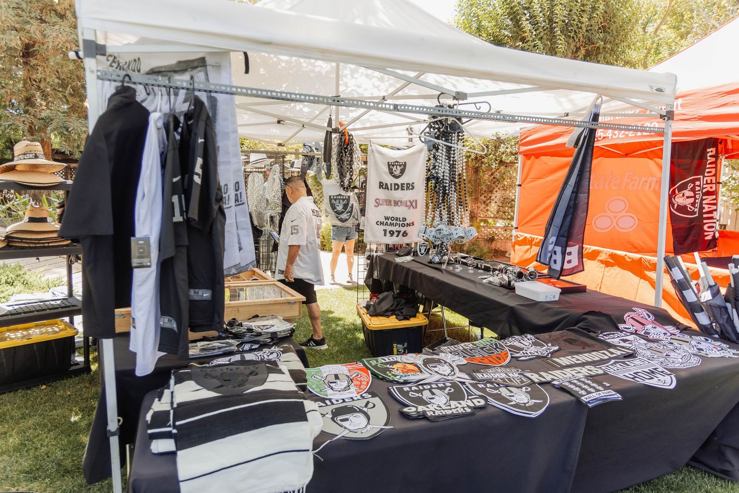 Outdoor Raiders NFL merchandise booth displaying apparel, patches, and accessories under white and orange canopy tents.