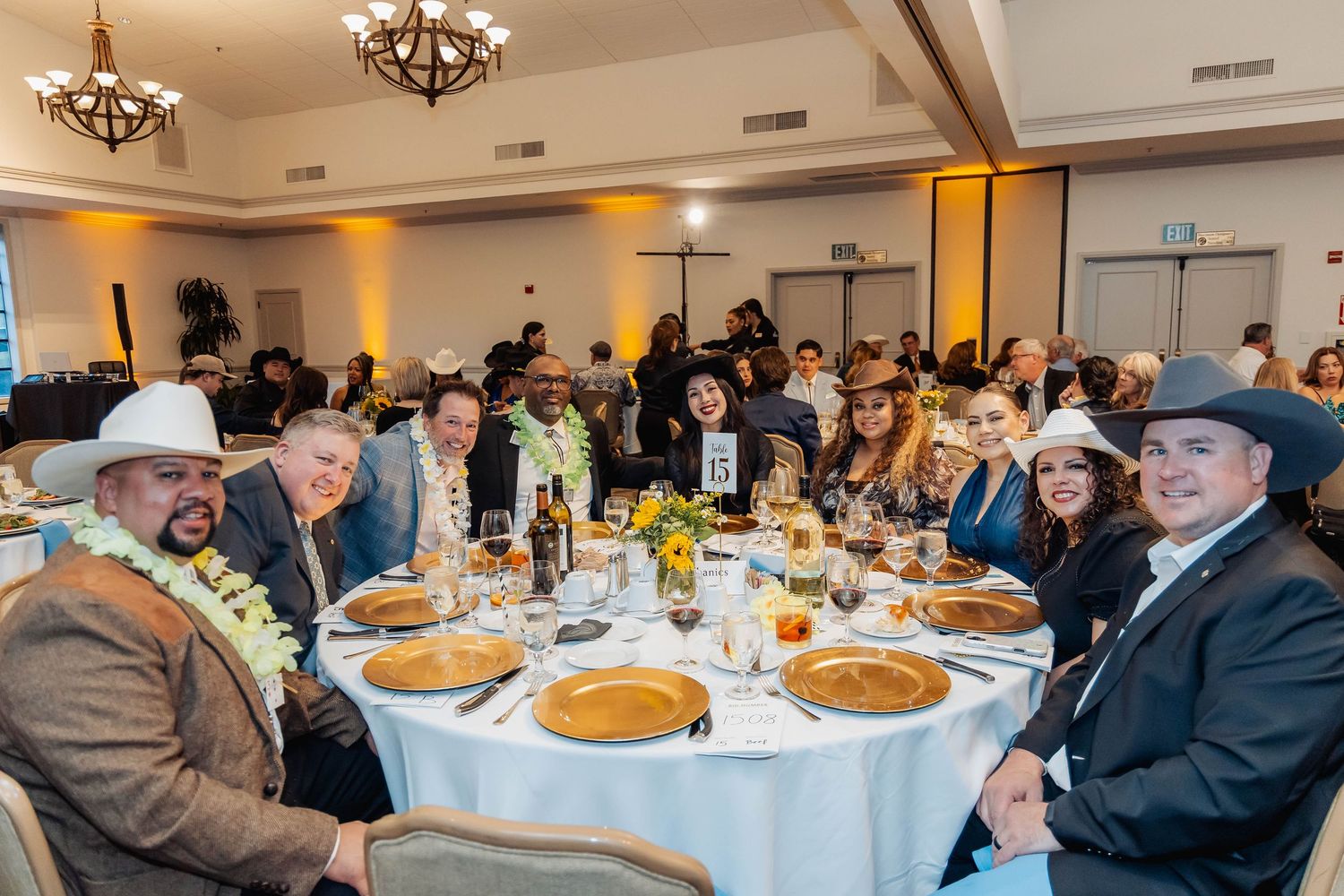 Elegantly dressed guests smile at a round banquet table during a formal gala event in a warmly lit ballroom.