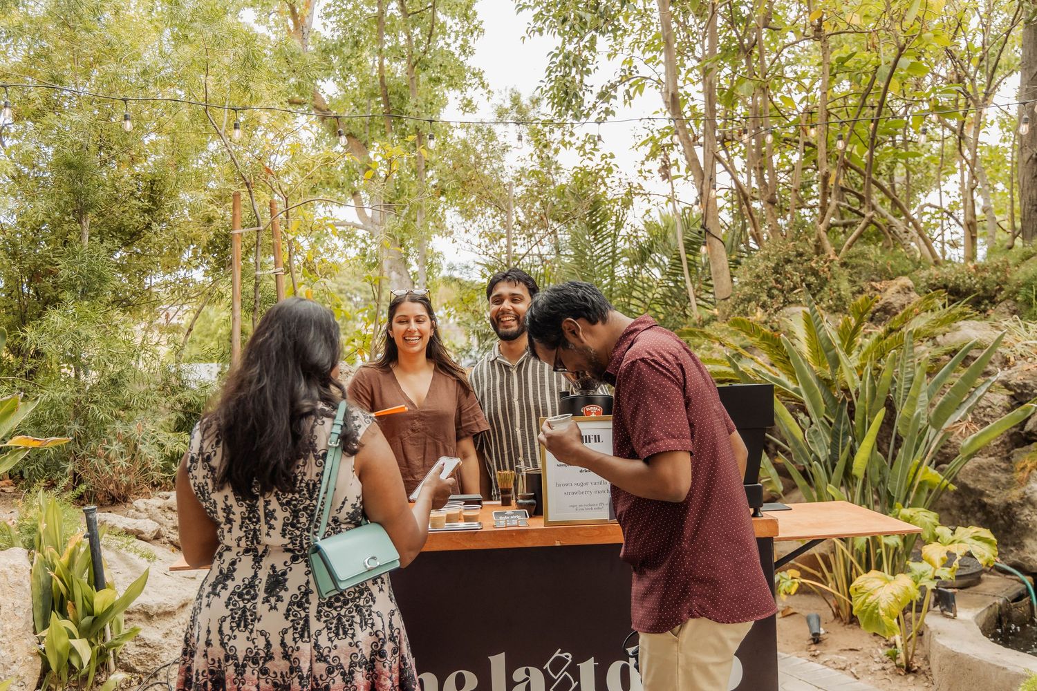 Four friends gather around an outdoor bar stand surrounded by lush green trees at a garden event.