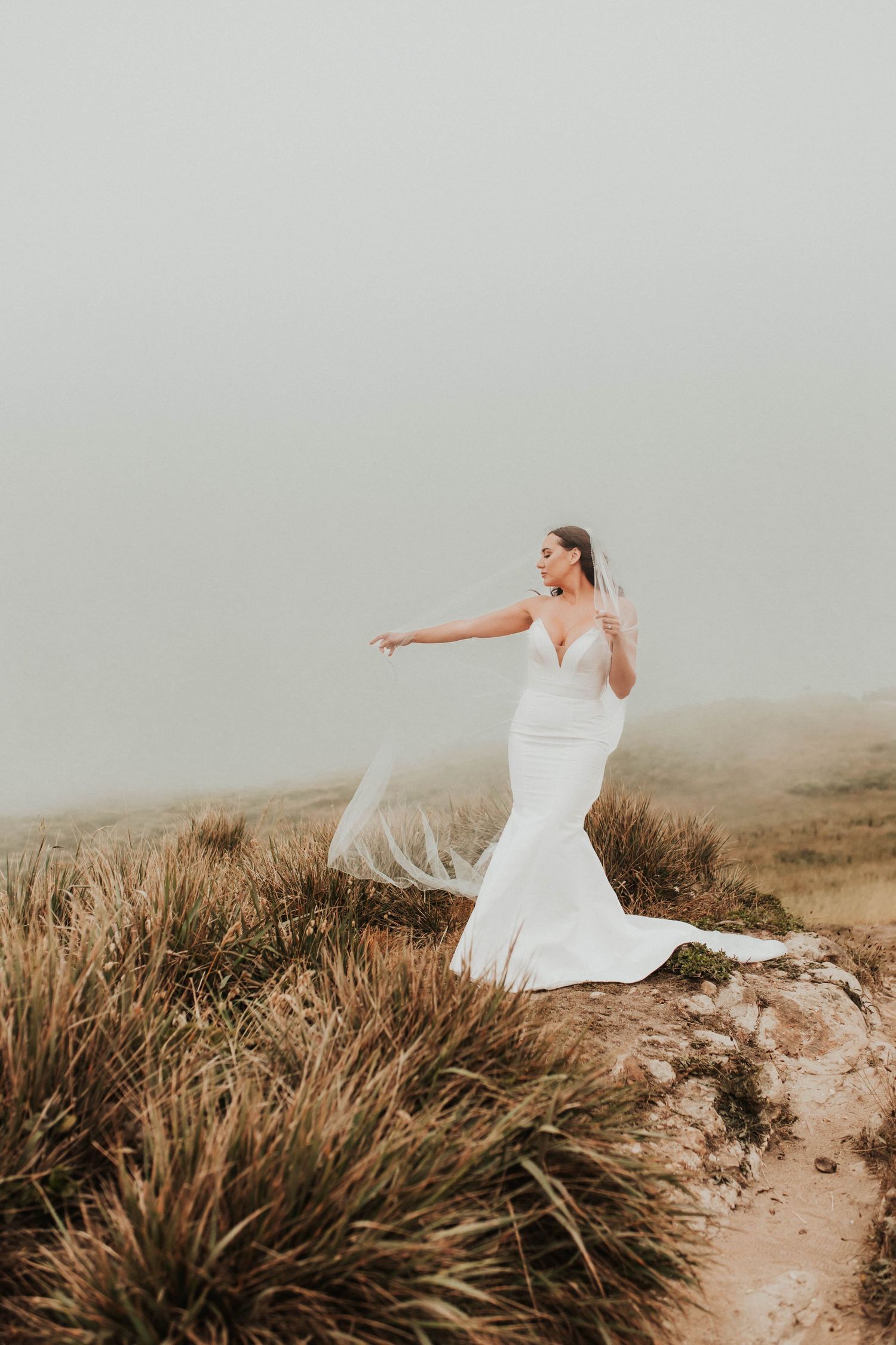A bride in a fitted white wedding dress poses dramatically on a misty hilltop with tall grass.