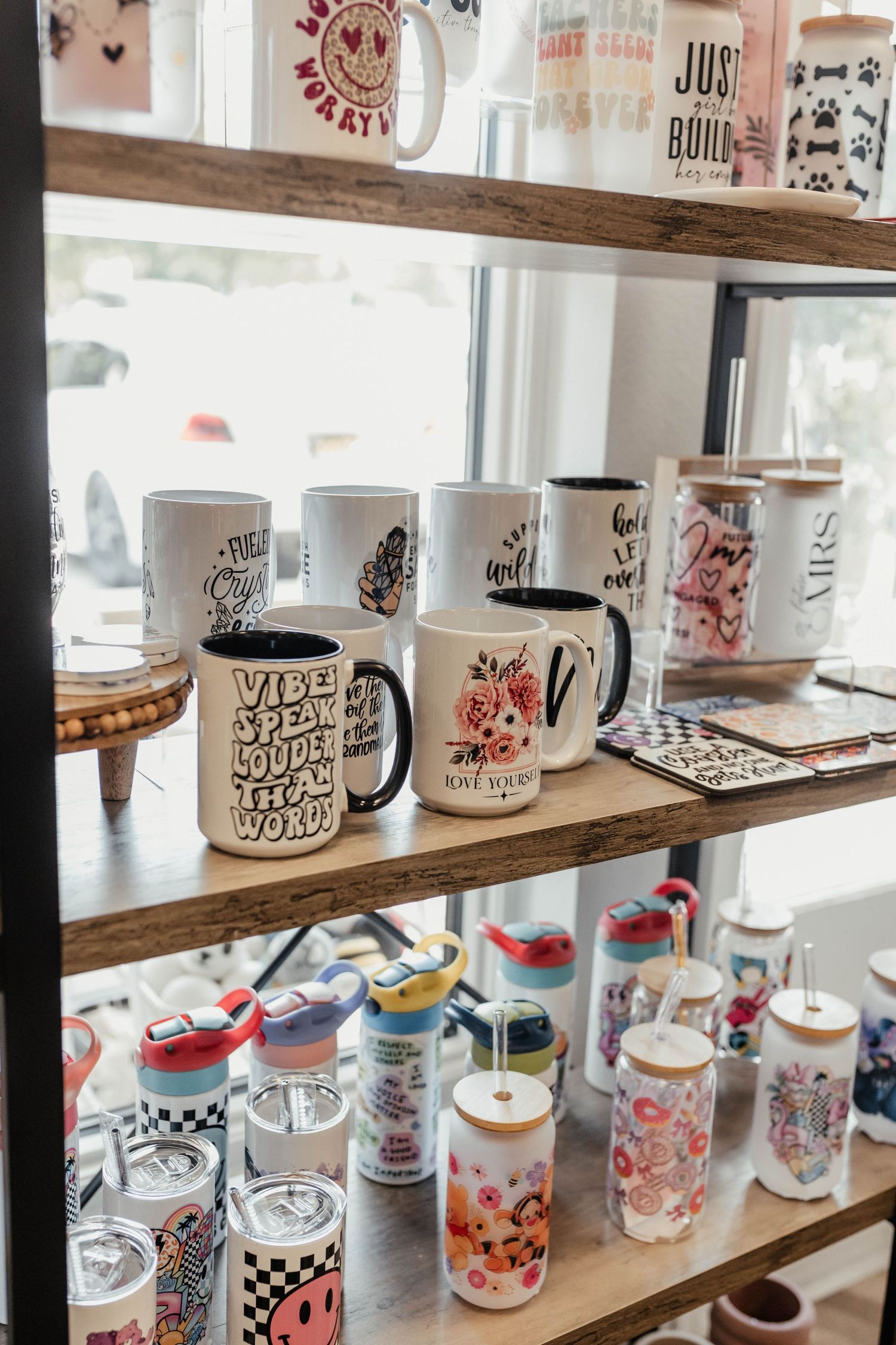 Collection of decorative mugs and tumblers with artistic designs displayed on wooden retail shelves.