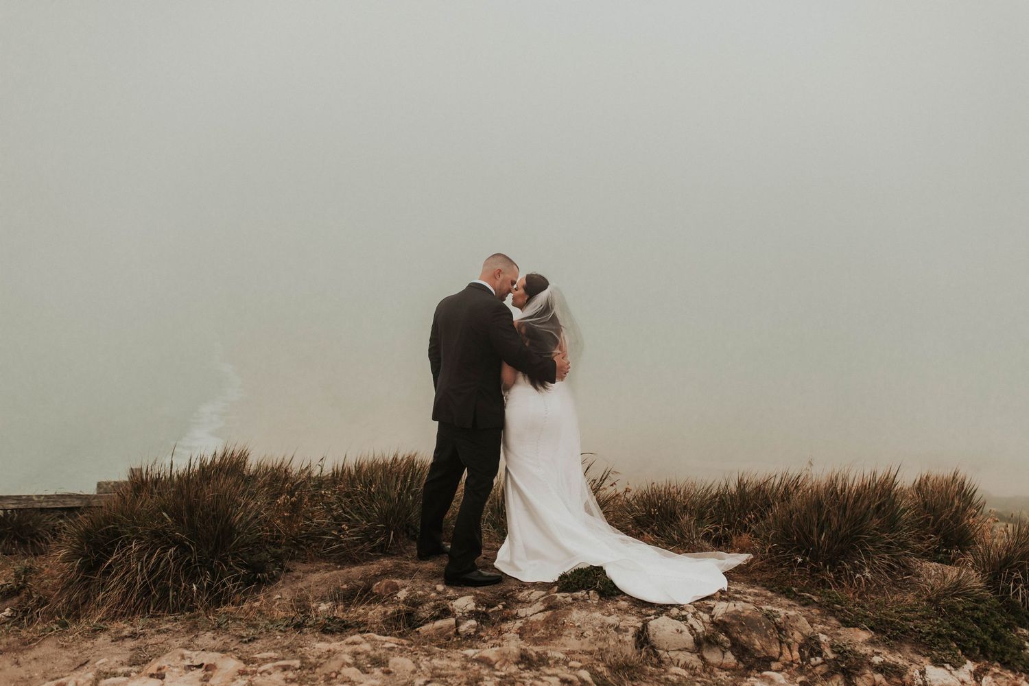 Newlyweds embrace on a foggy clifftop with dried grass surrounding them.