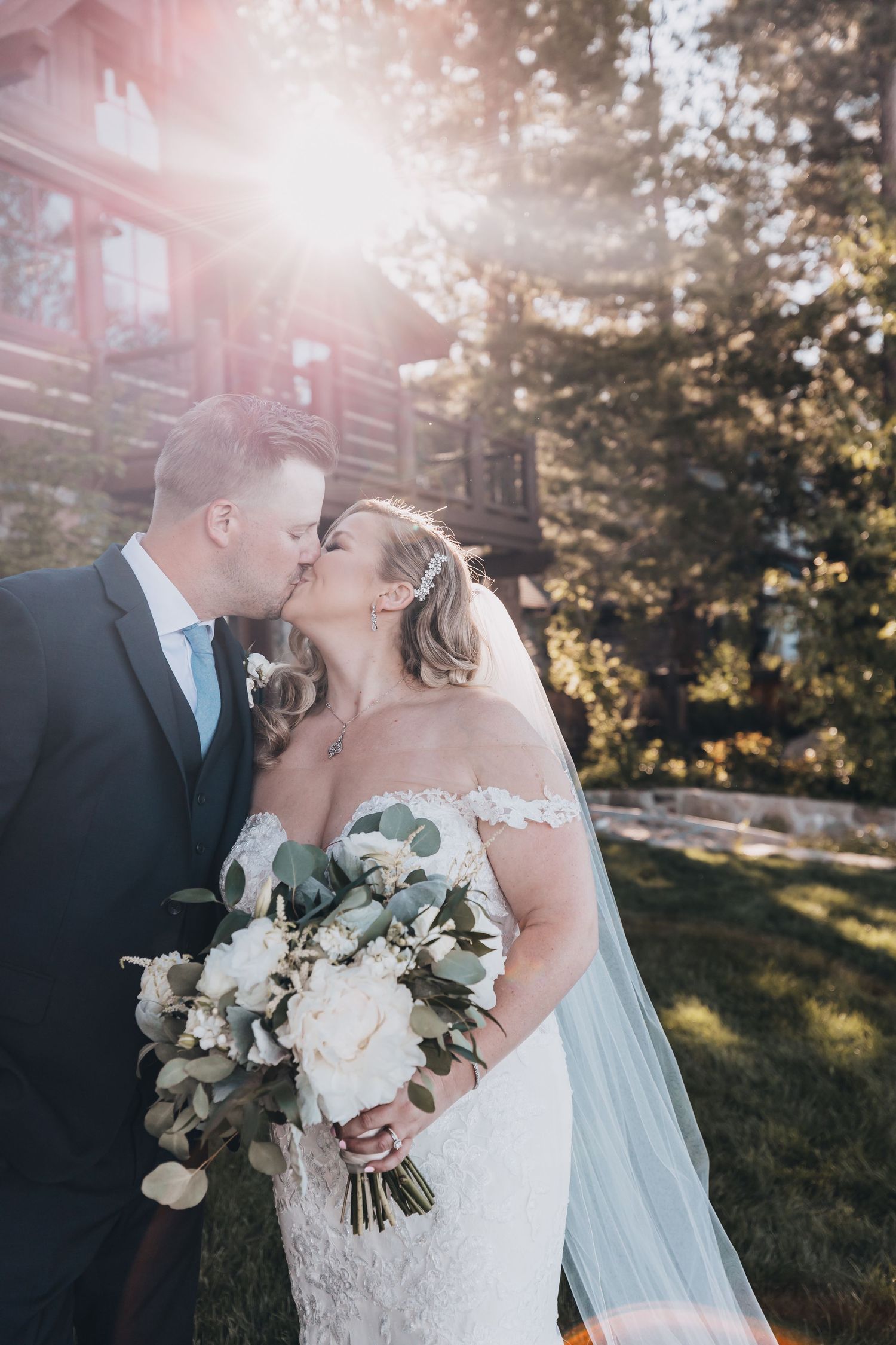 Bride holding white floral bouquet shares romantic moment with groom in sunlit outdoor setting.