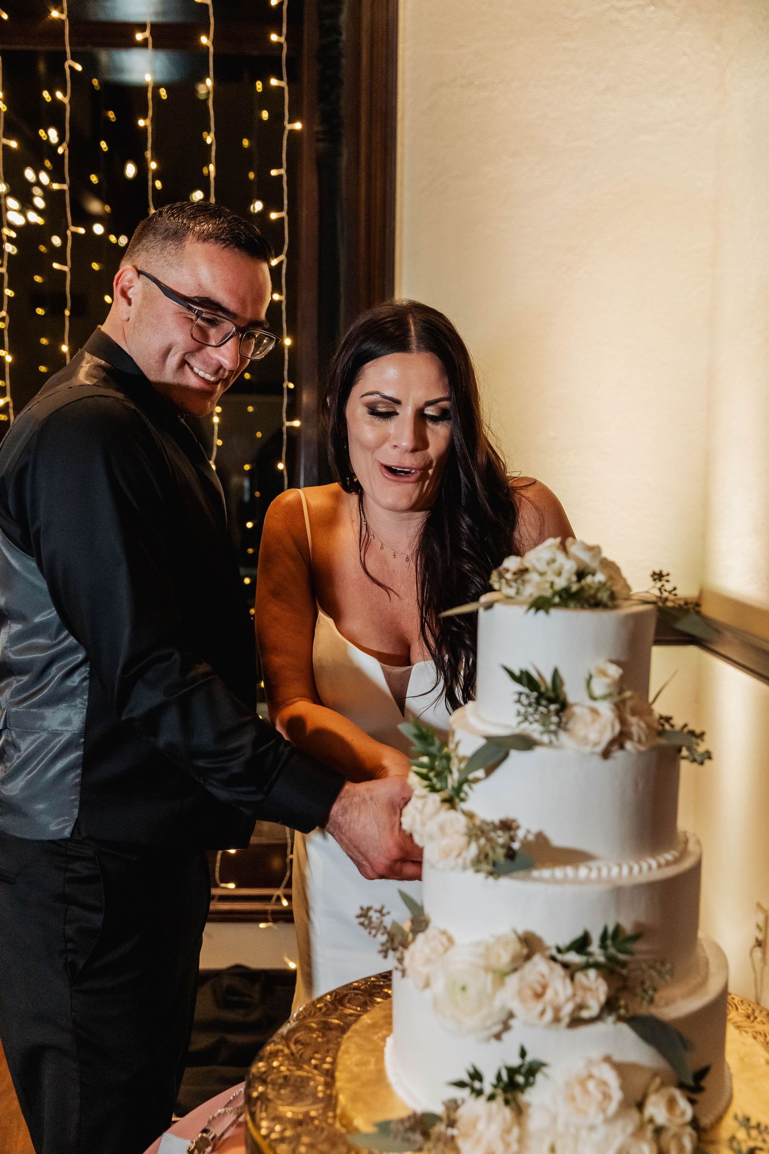 Wedding couple cuts their elegant white tiered cake decorated with flowers and greenery.