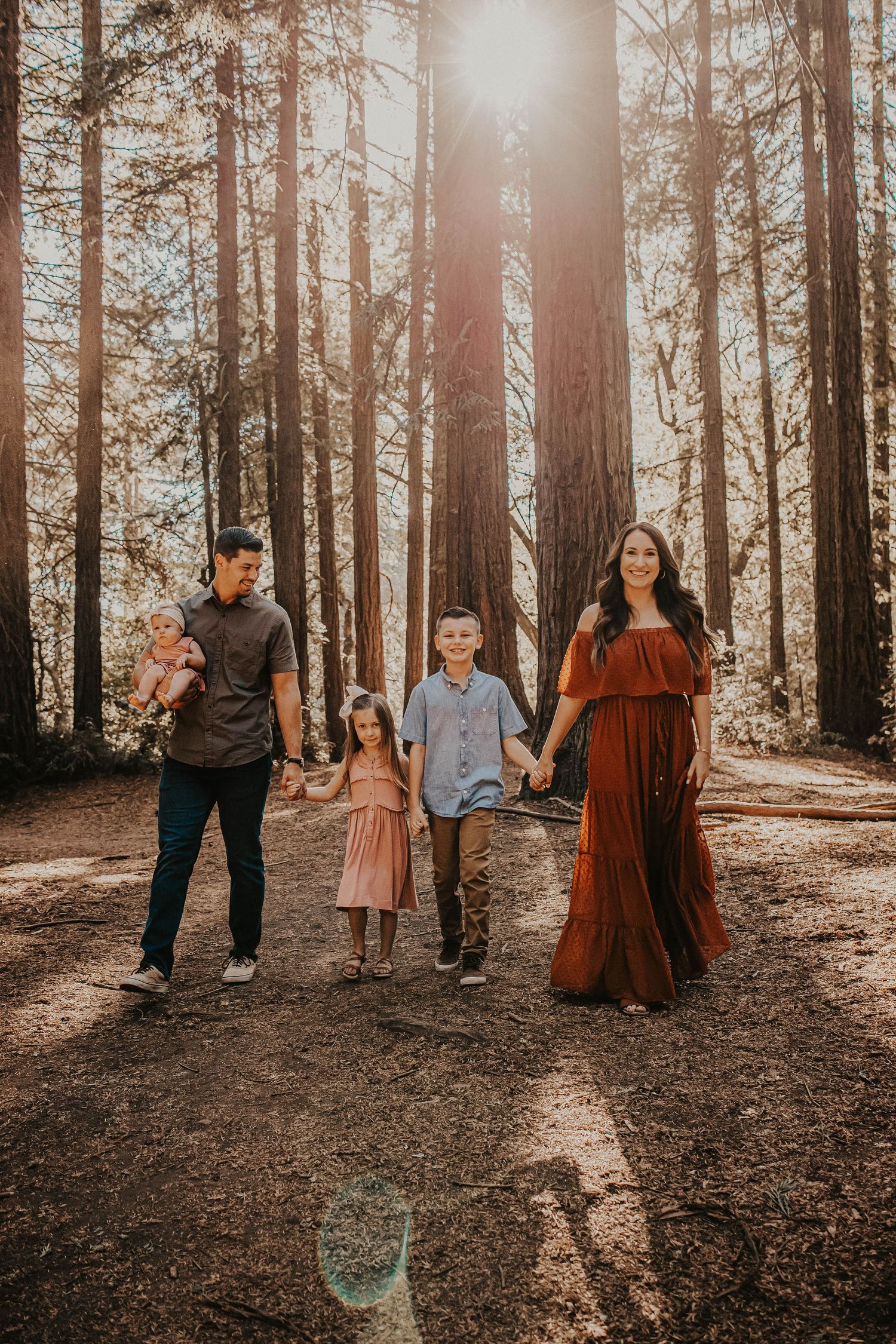 Family of five walking through a sunlit redwood forest, parents holding hands with children in warm autumn tones.