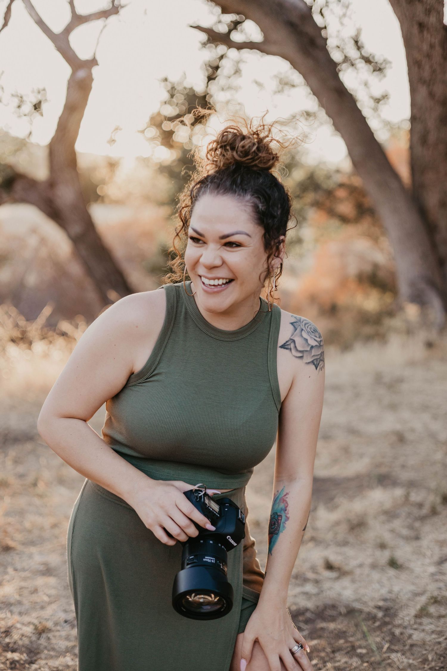 A photographer in an olive green dress laughs while holding a professional camera outdoors during golden hour.