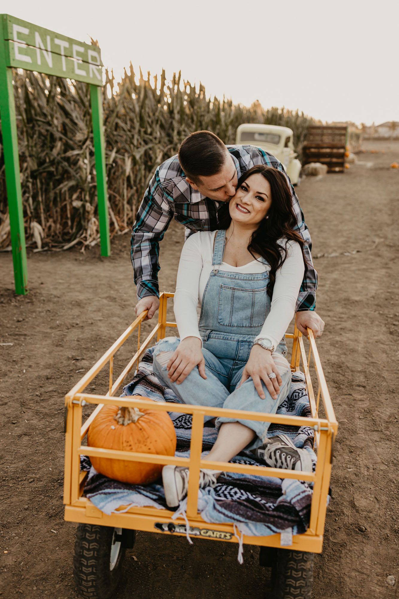 A couple poses playfully in a wooden wagon with pumpkins and blankets at a fall farm.