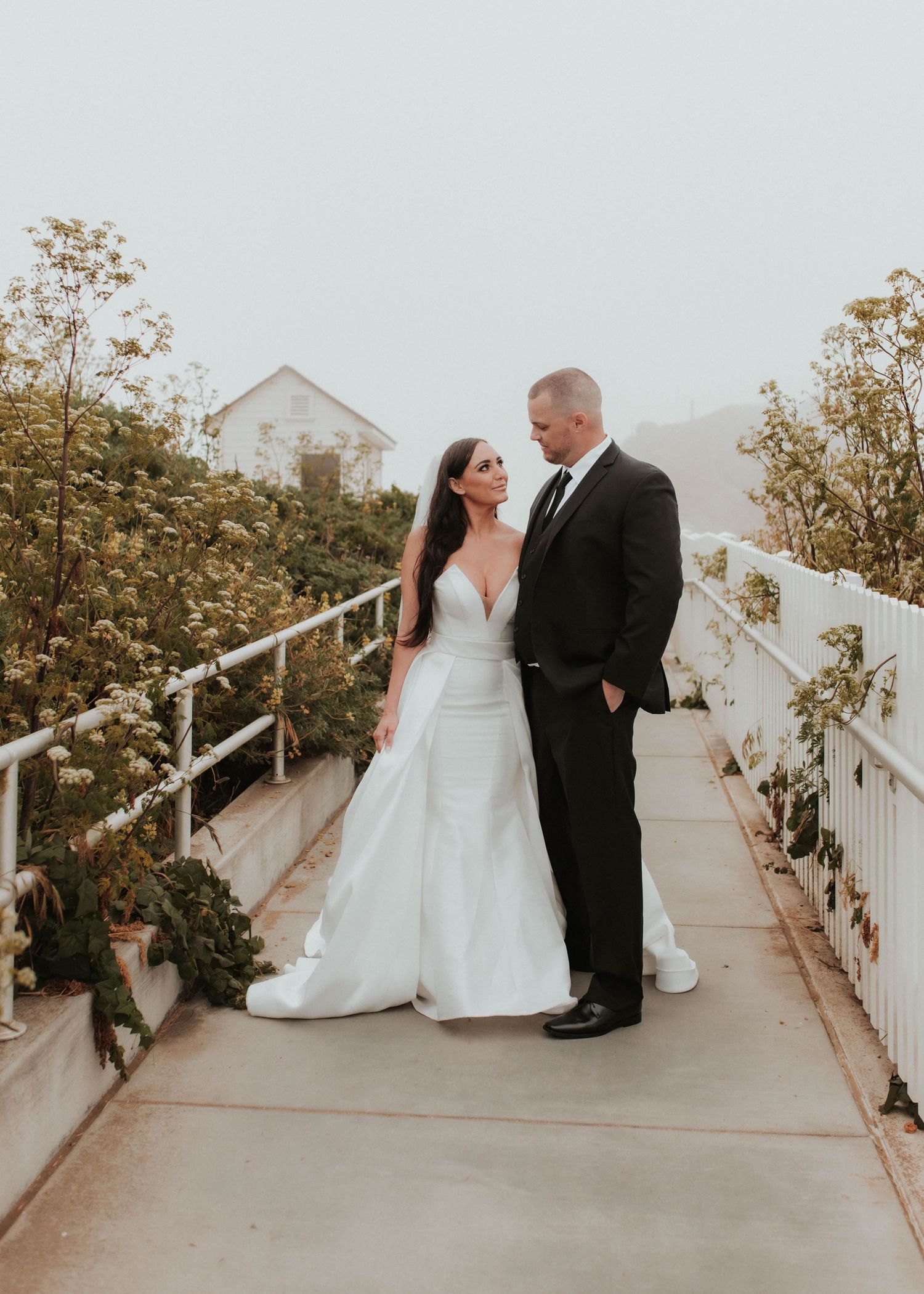 Wedding couple stands together on garden path with white picket fence and house in background.