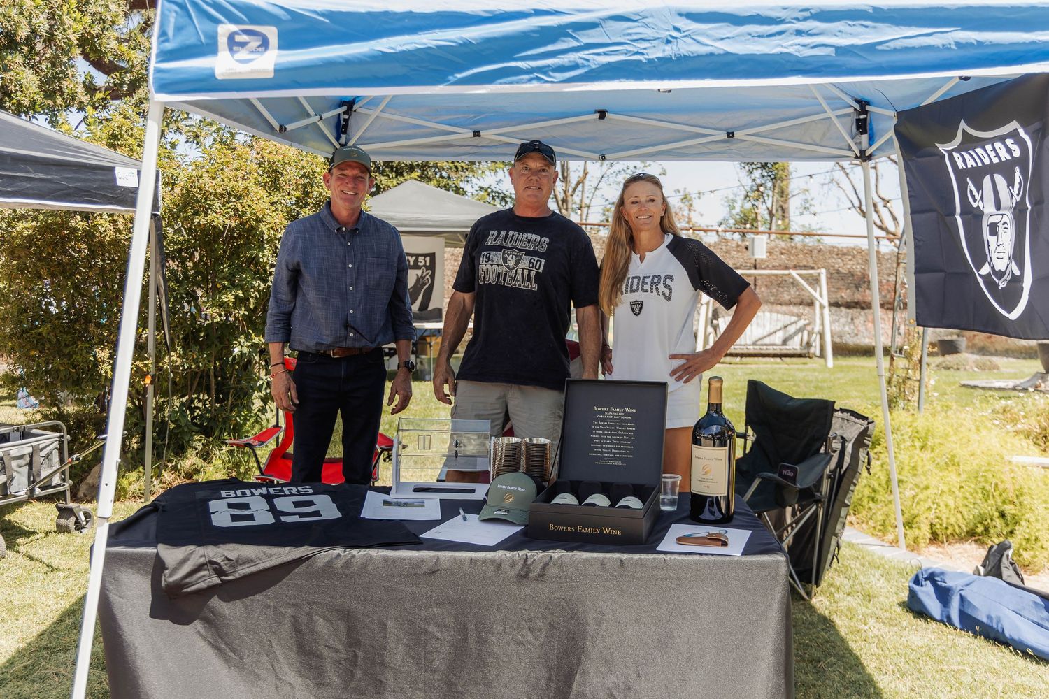 Three people standing behind a Raiders-themed vendor booth under a blue canopy tent at an outdoor event.