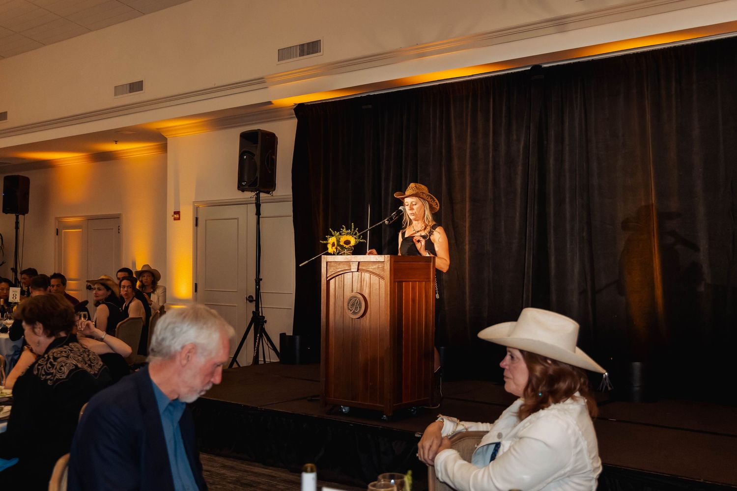 Woman speaking at podium on stage at formal event, audience seated at tables, warm amber lighting.