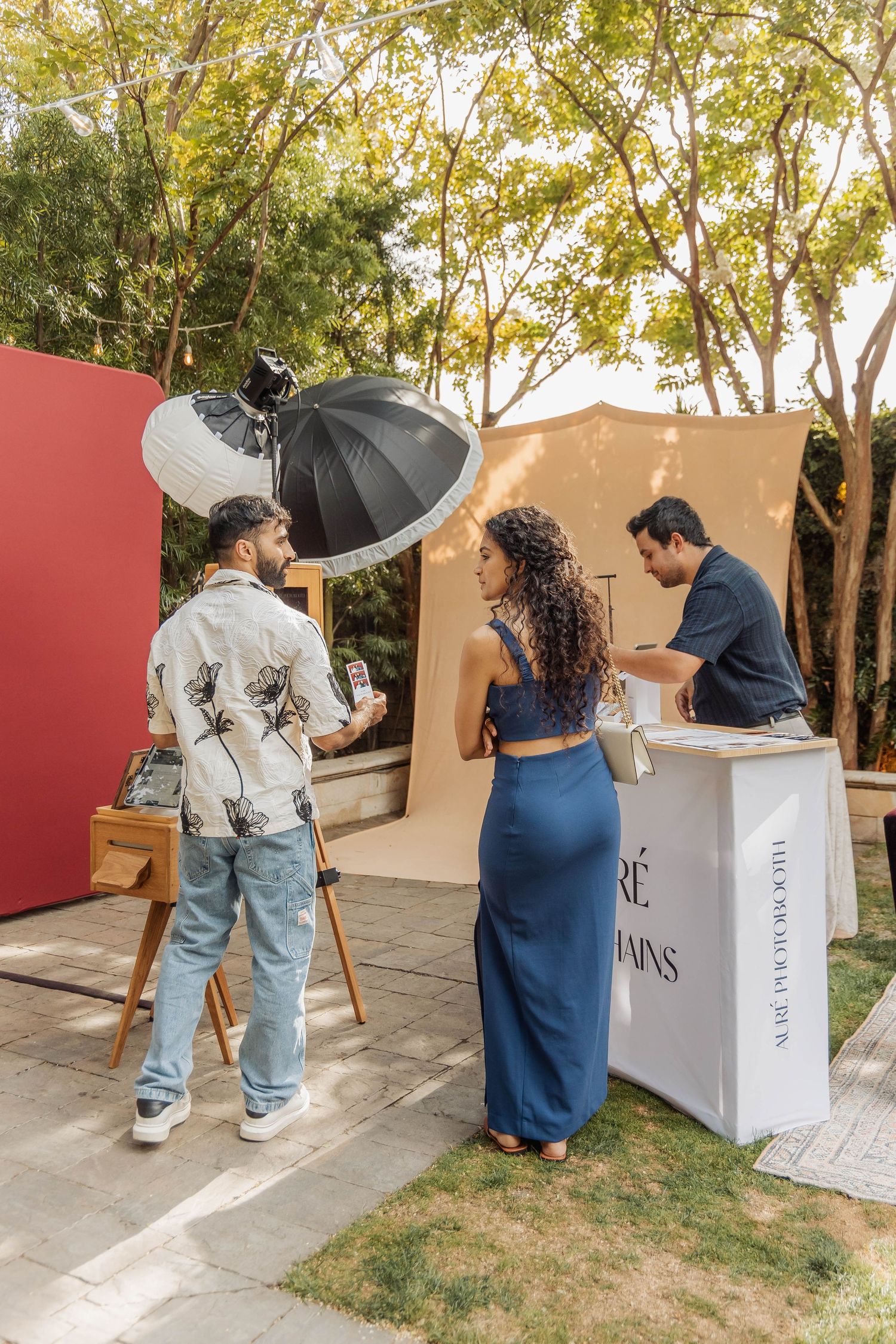 Outdoor photo booth setup with studio lighting, two guests posing near a branded white podium surrounded by trees.