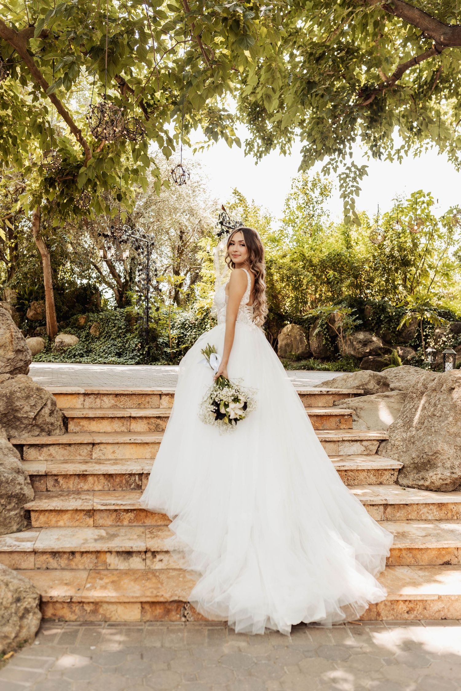 A wedding dress with flowing tulle skirt photographed on stone steps in a garden setting with natural lighting.