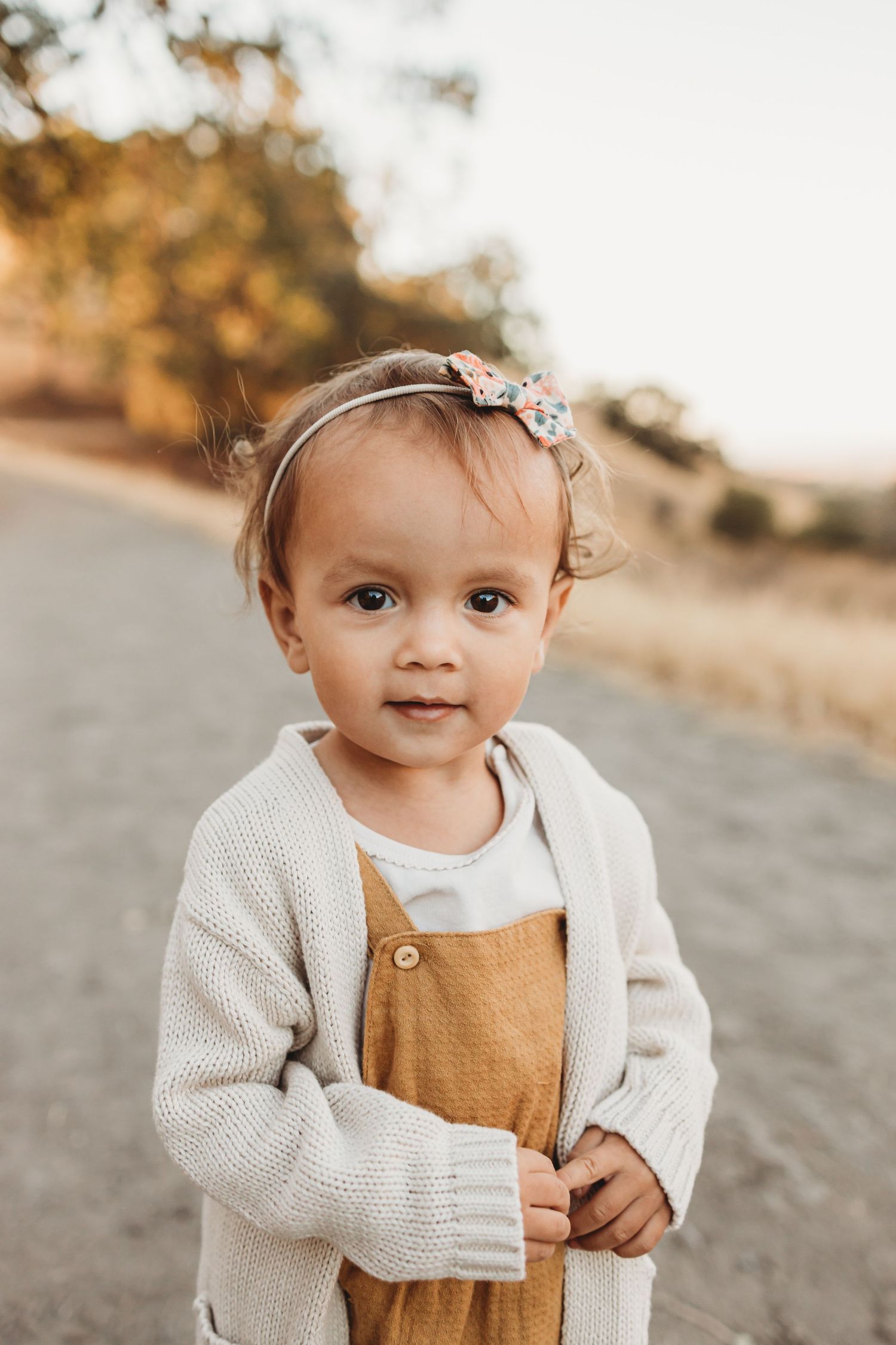 Toddler wearing cream cardigan and brown overalls smiling on outdoor trail.
