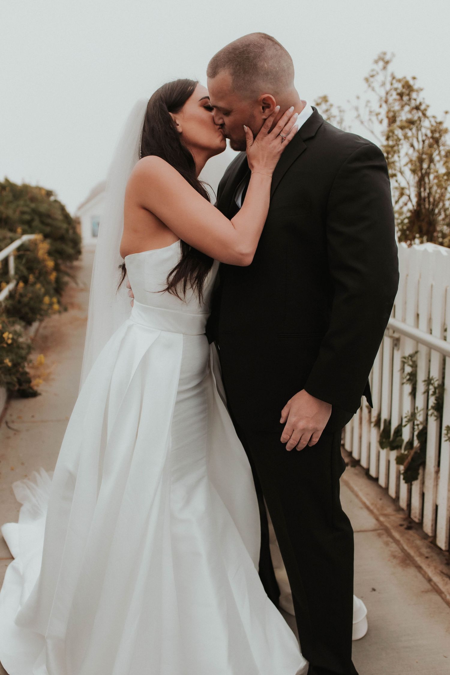 Romantic kiss between newlyweds on garden pathway with white fence.