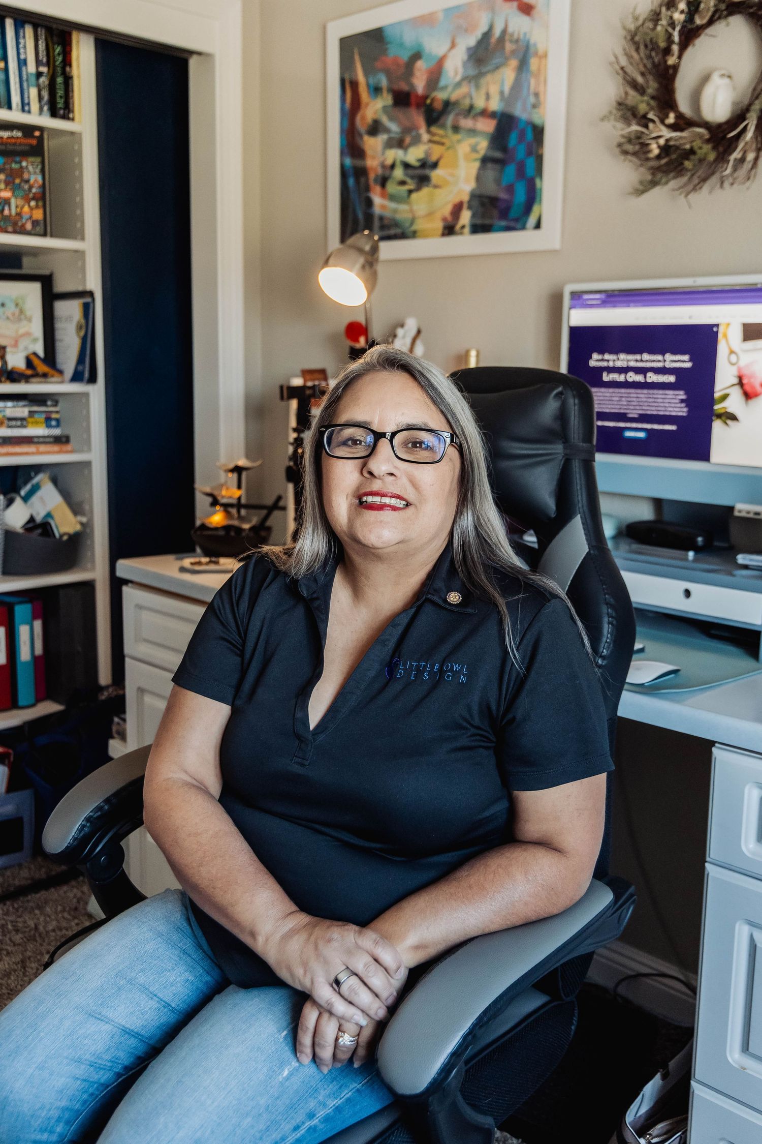 Person in black polo shirt sitting at home office desk with computer monitors and decorative wall art.