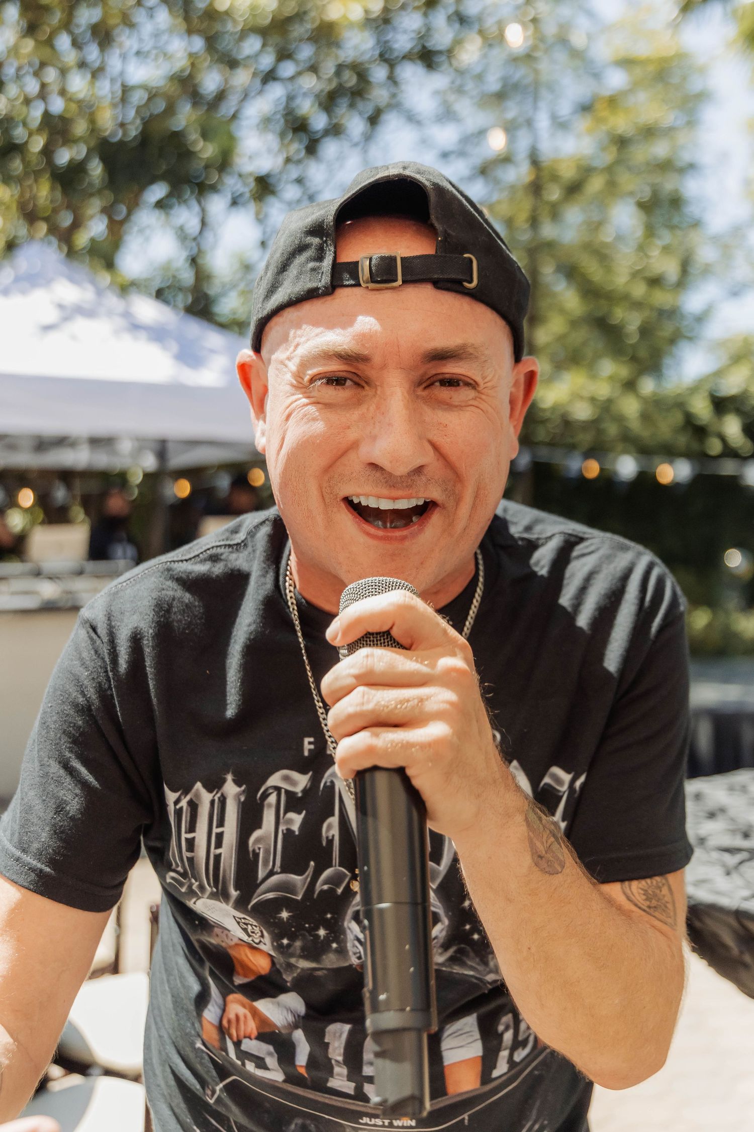 Smiling man in black cap holding microphone performing at outdoor Raiders themed event.