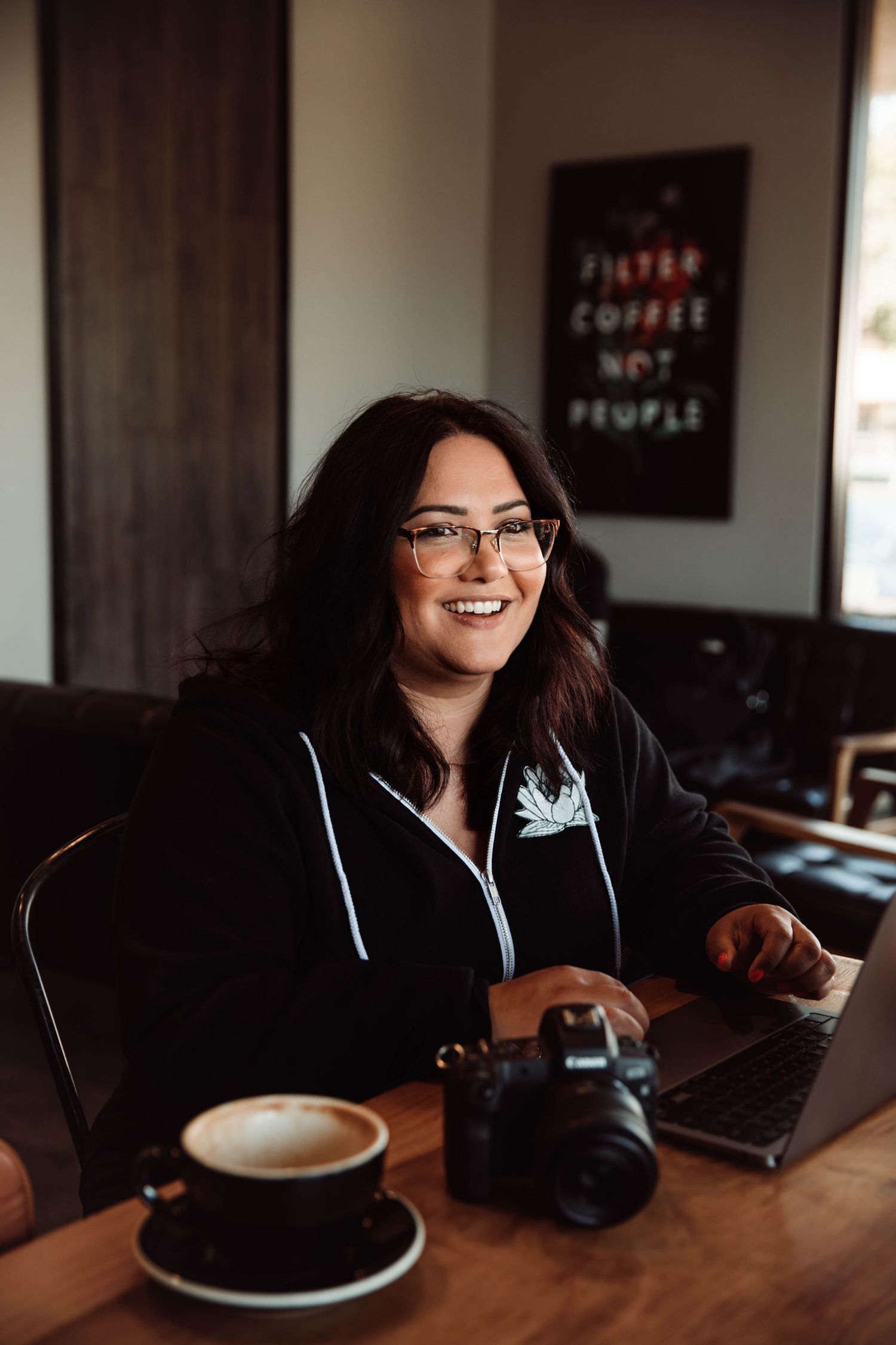 Smiling woman with glasses working on laptop at cafe table with coffee and camera, poster reading Filter Coffee Not People behind her.
