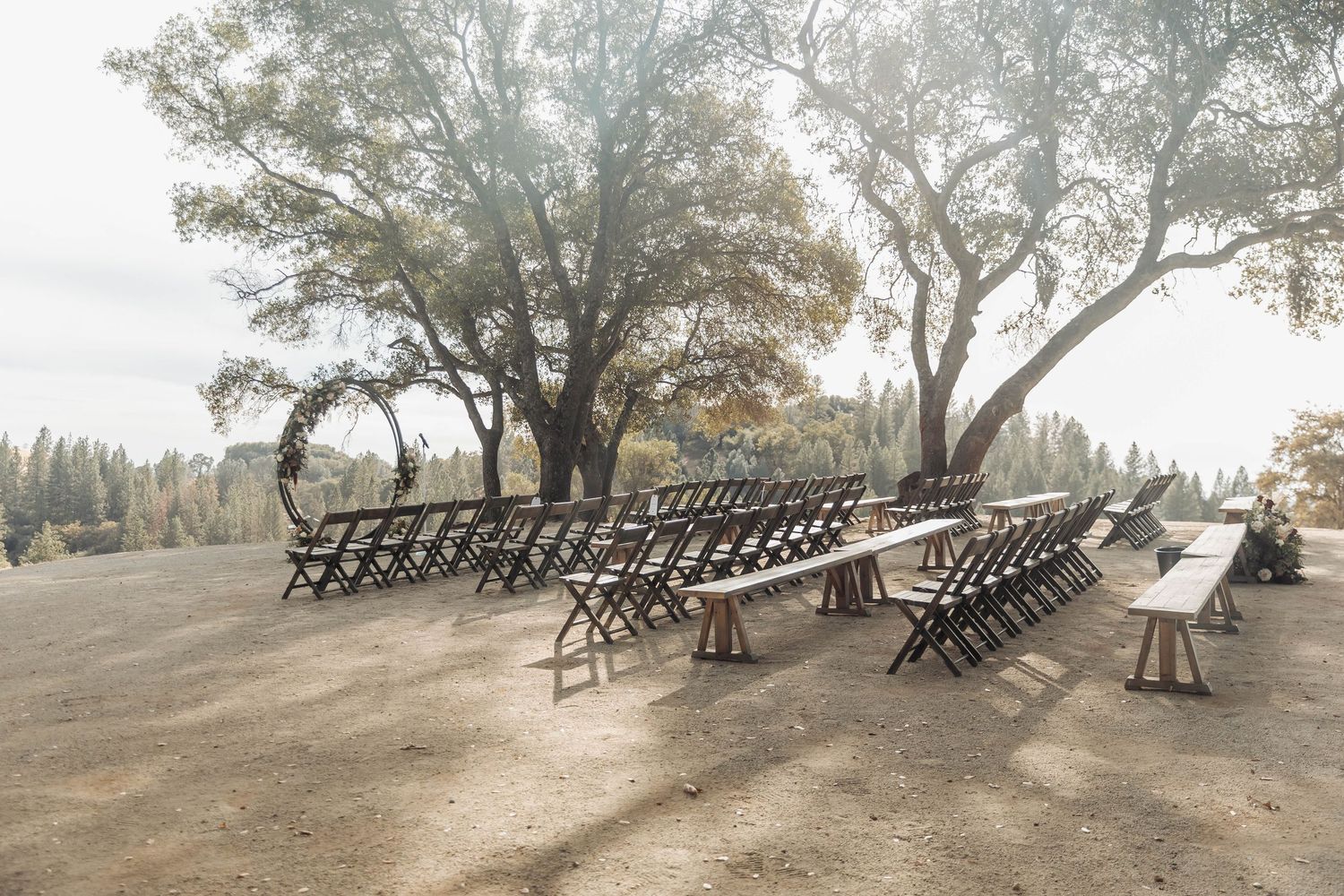 Rustic outdoor wedding ceremony setup with wooden chairs under large trees.