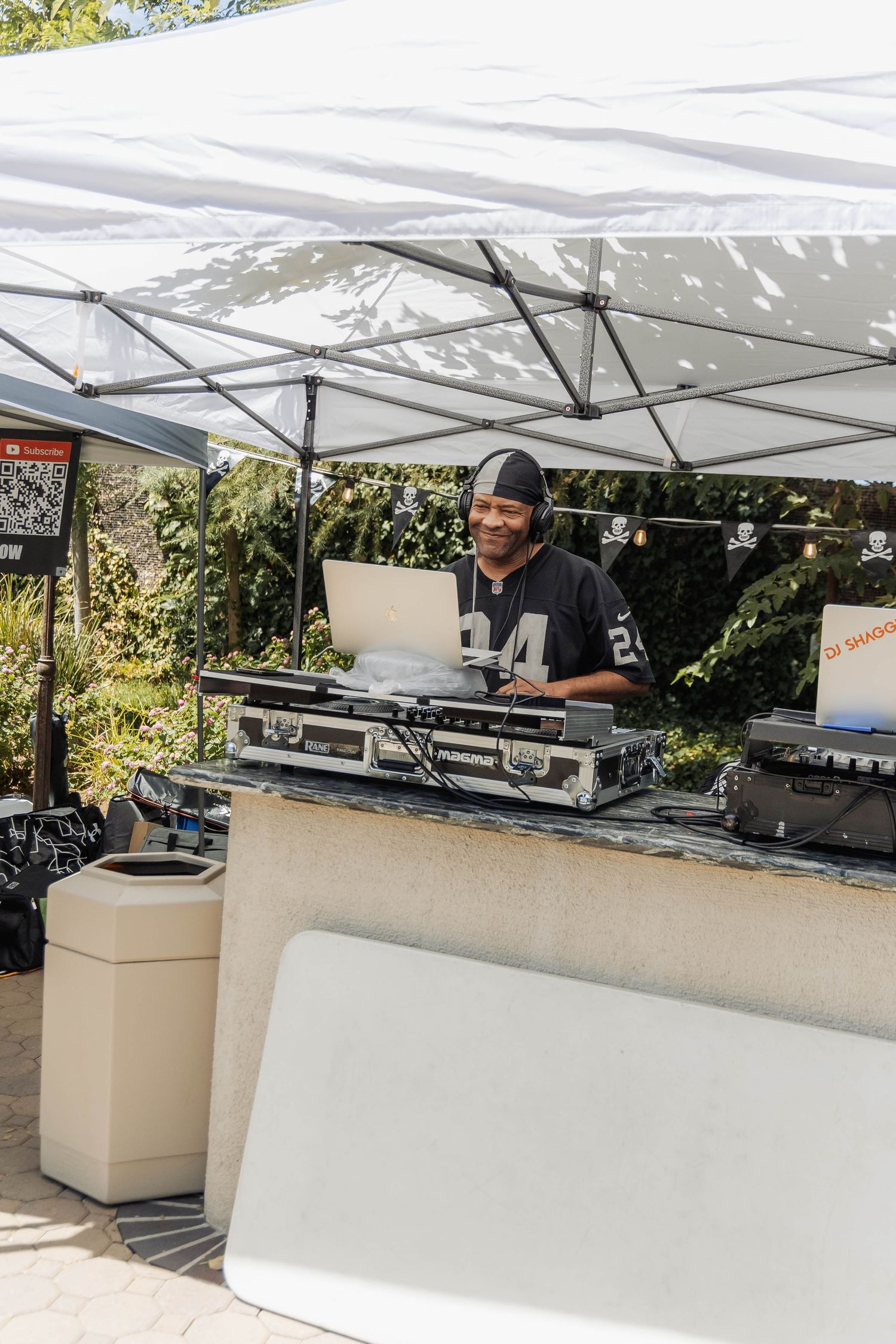Smiling DJ in Raiders jersey operating turntables and laptop under white canopy at outdoor event.