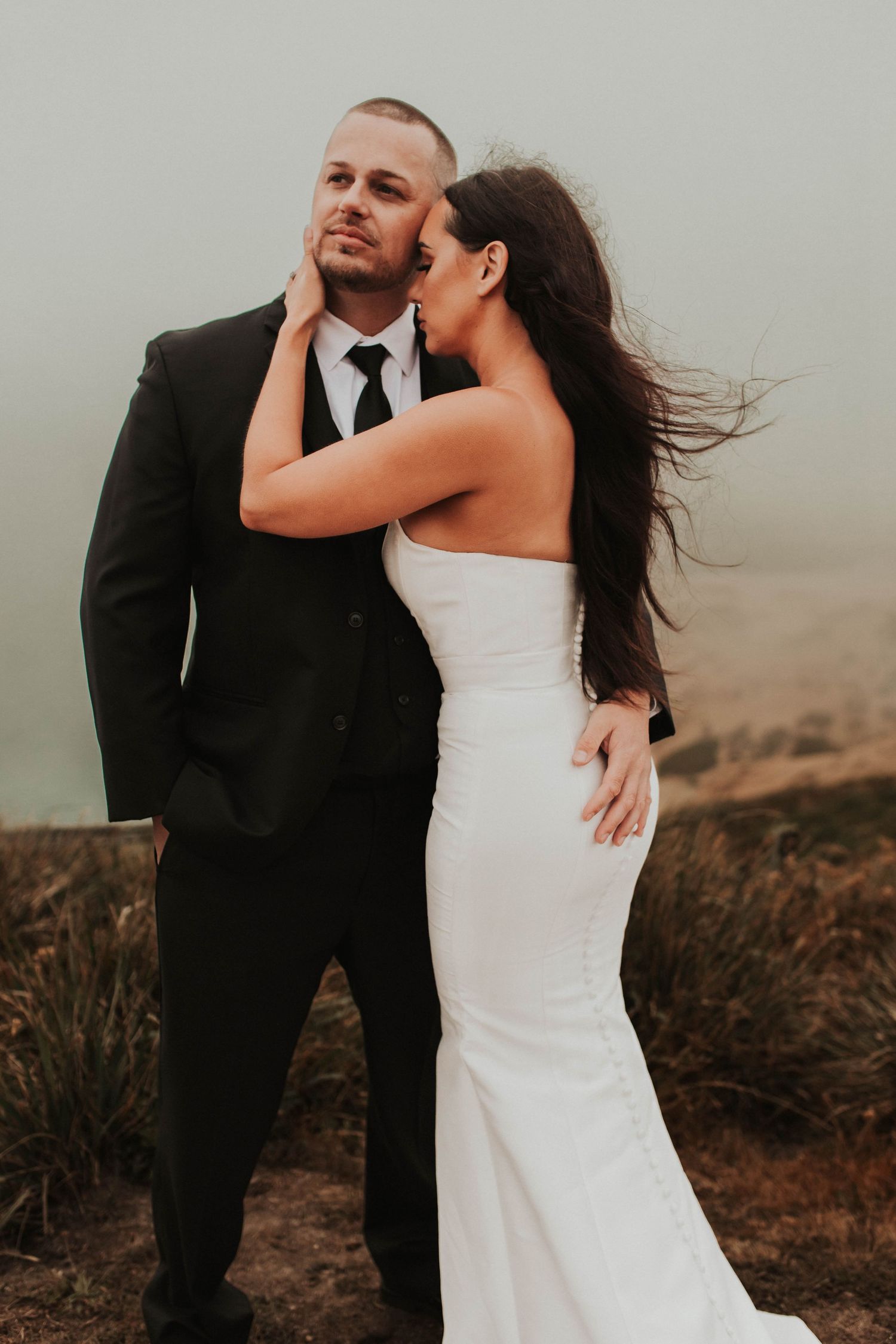 An intimate wedding portrait captured on a windswept hilltop at dusk.