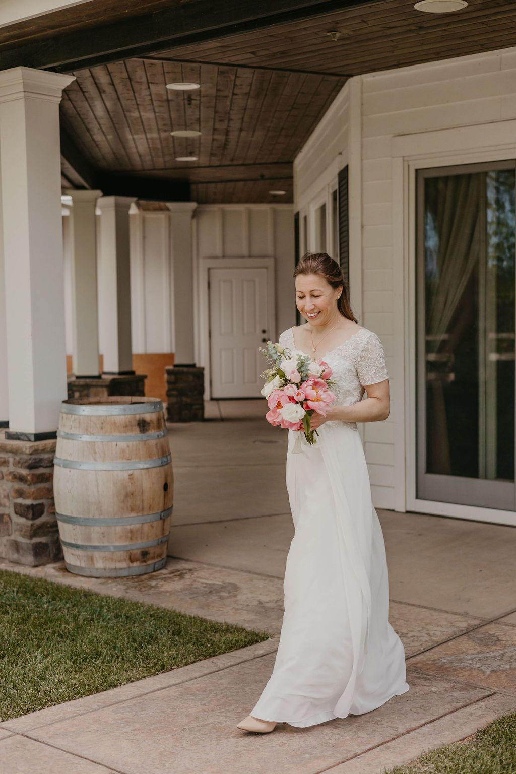 A person in a white wedding dress holds a pink bouquet while walking on a covered porch.