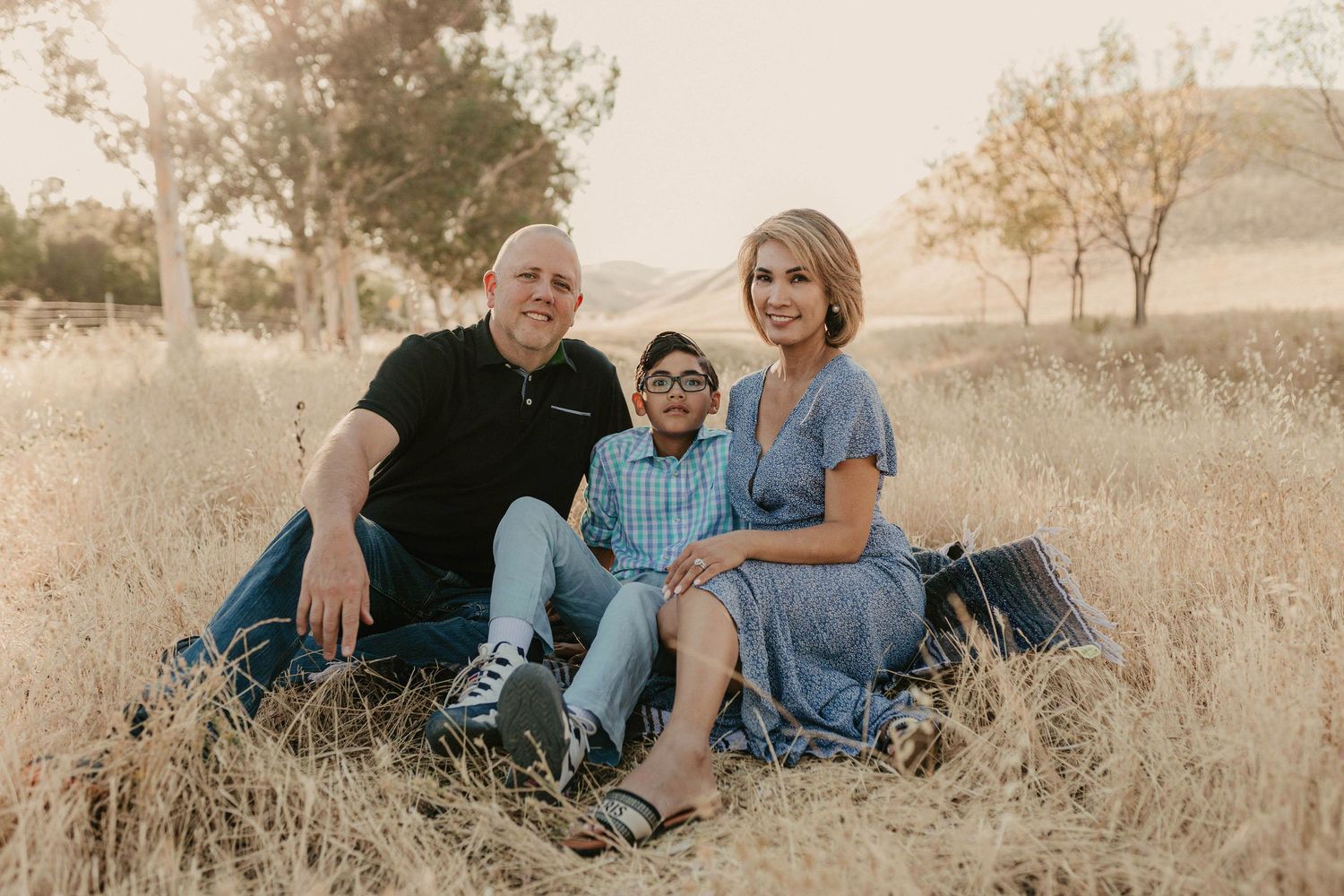 Family of three smiling outdoors in golden field, parents sitting with young boy in glasses during sunset photo session.