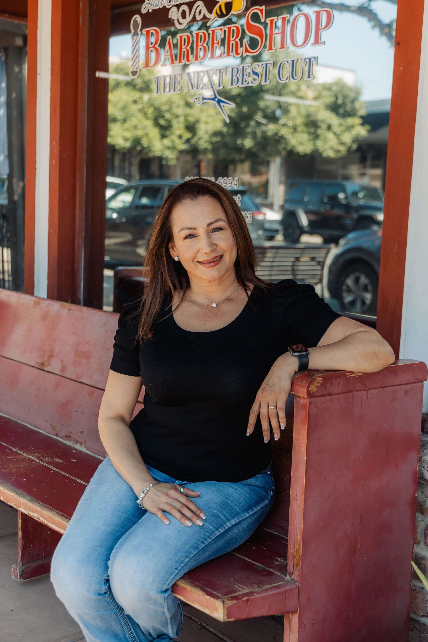 Portrait of someone in black top and jeans posed on red bench outside barber shop entrance.