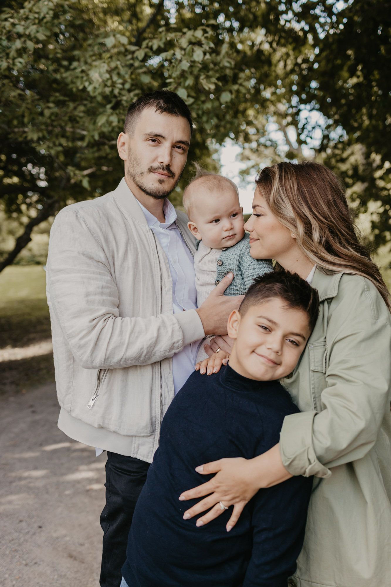 Beautiful family portrait taken outdoors with parents and two children in casual attire on a tree-lined path.