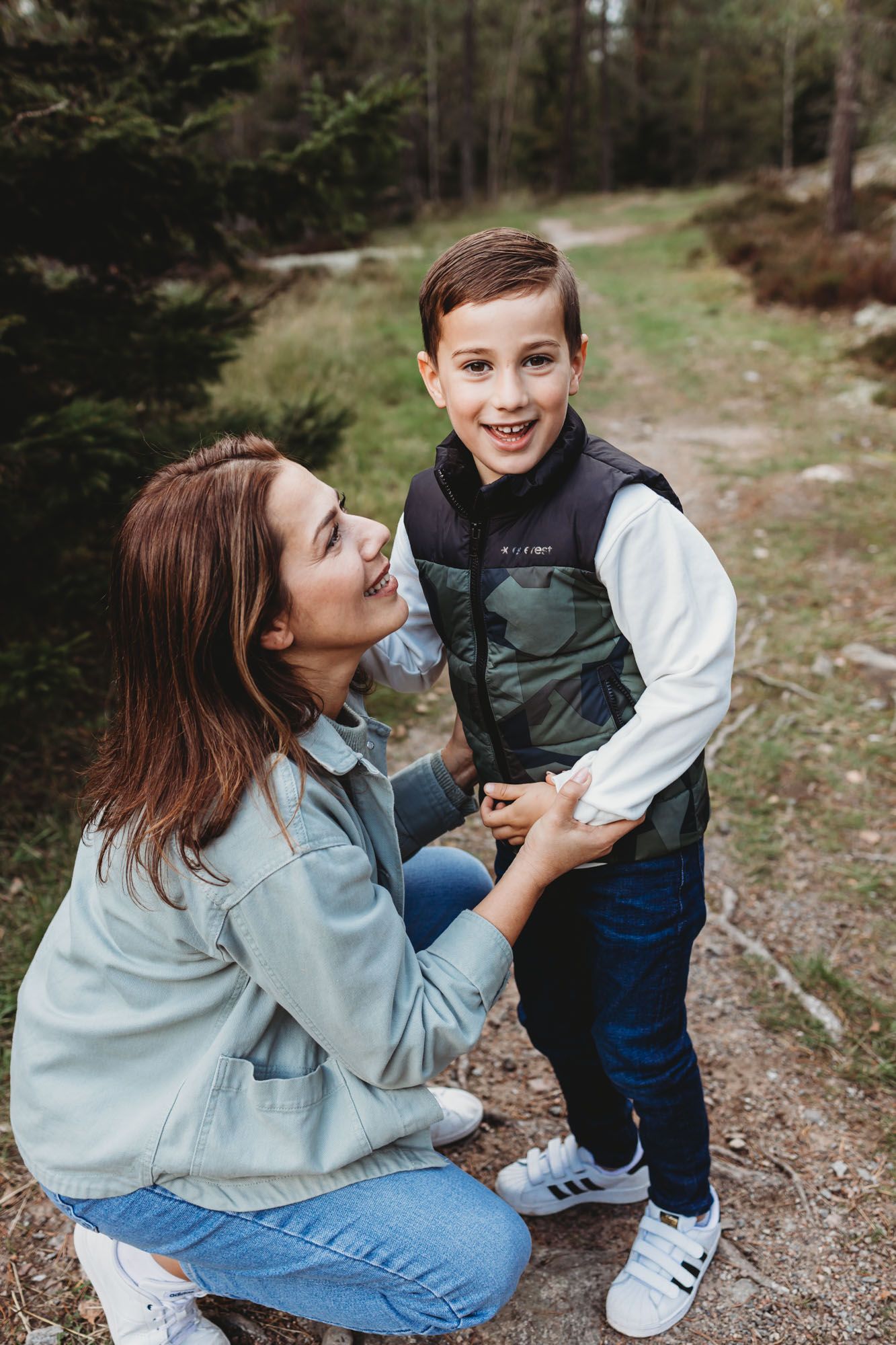 Young person in denim jacket kneeling beside small child on forest path.