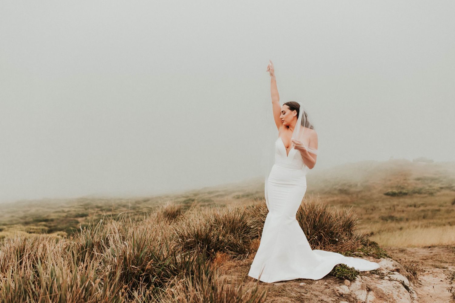 A bride stands on a hill with arms raised wearing a flowing mermaid wedding gown in foggy weather.