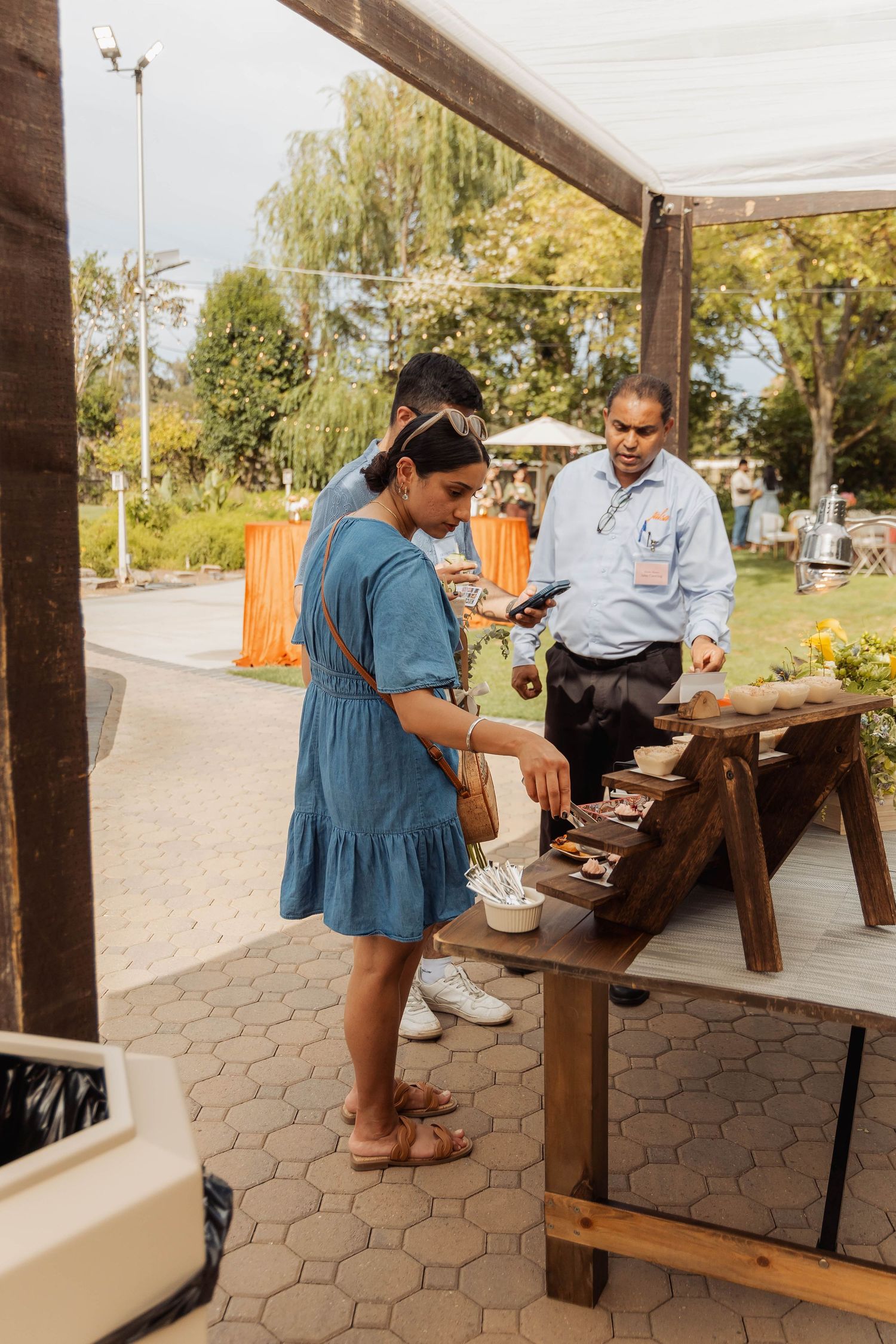 Woman in blue dress serving food at outdoor wooden buffet station during a garden event.