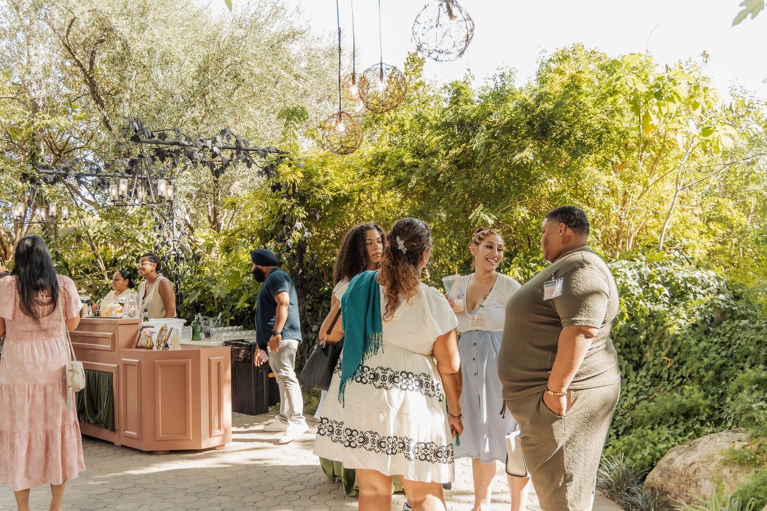 Groups of people socializing outdoors at a garden event surrounded by lush greenery and warm sunlight.